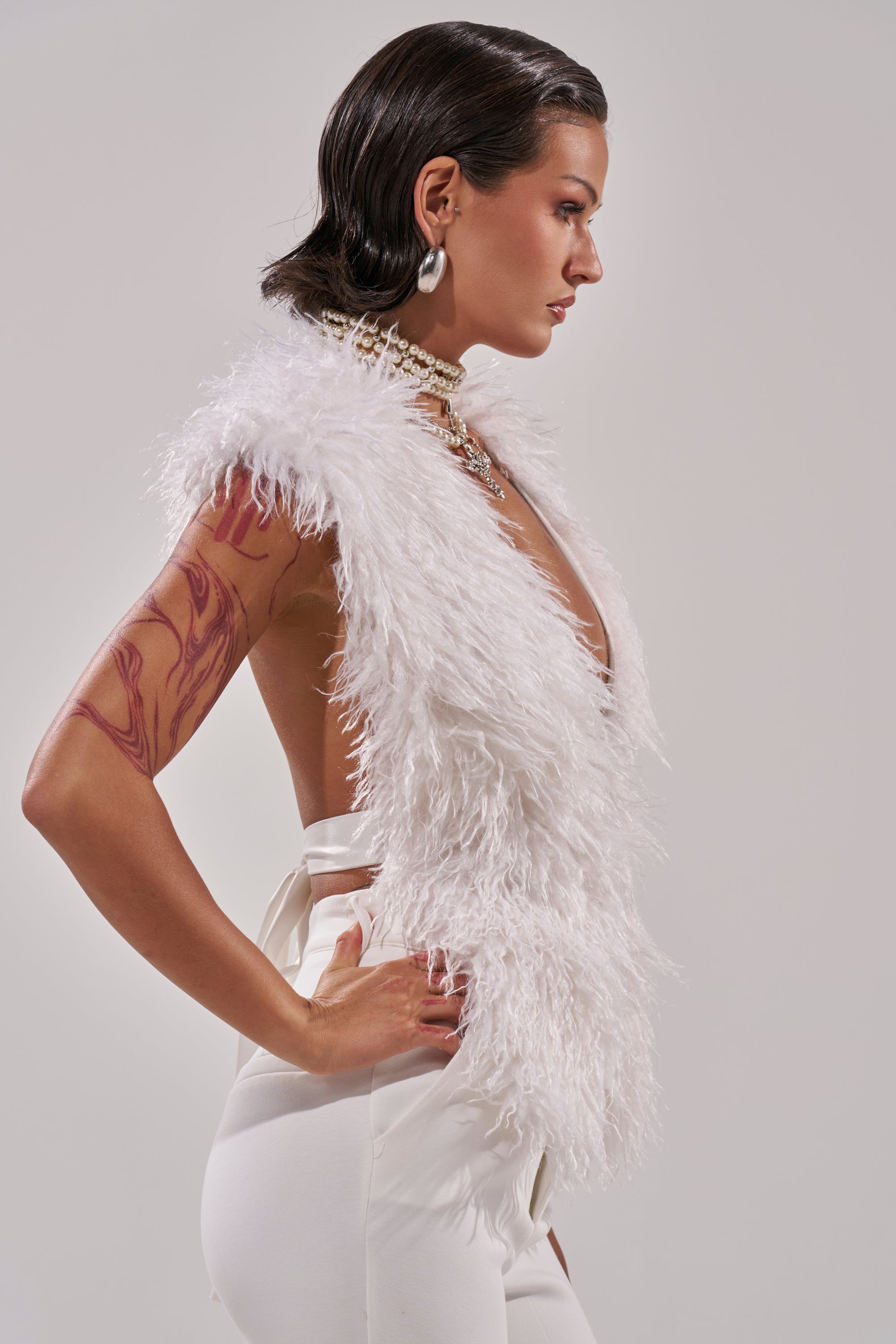 A woman in profile poses against a plain backdrop, wearing the ivory JAMEELA FAUX FUR TOP and high-waisted white pants. She has dark hair, statement earrings, layered necklaces, and a visible arm tattoo.