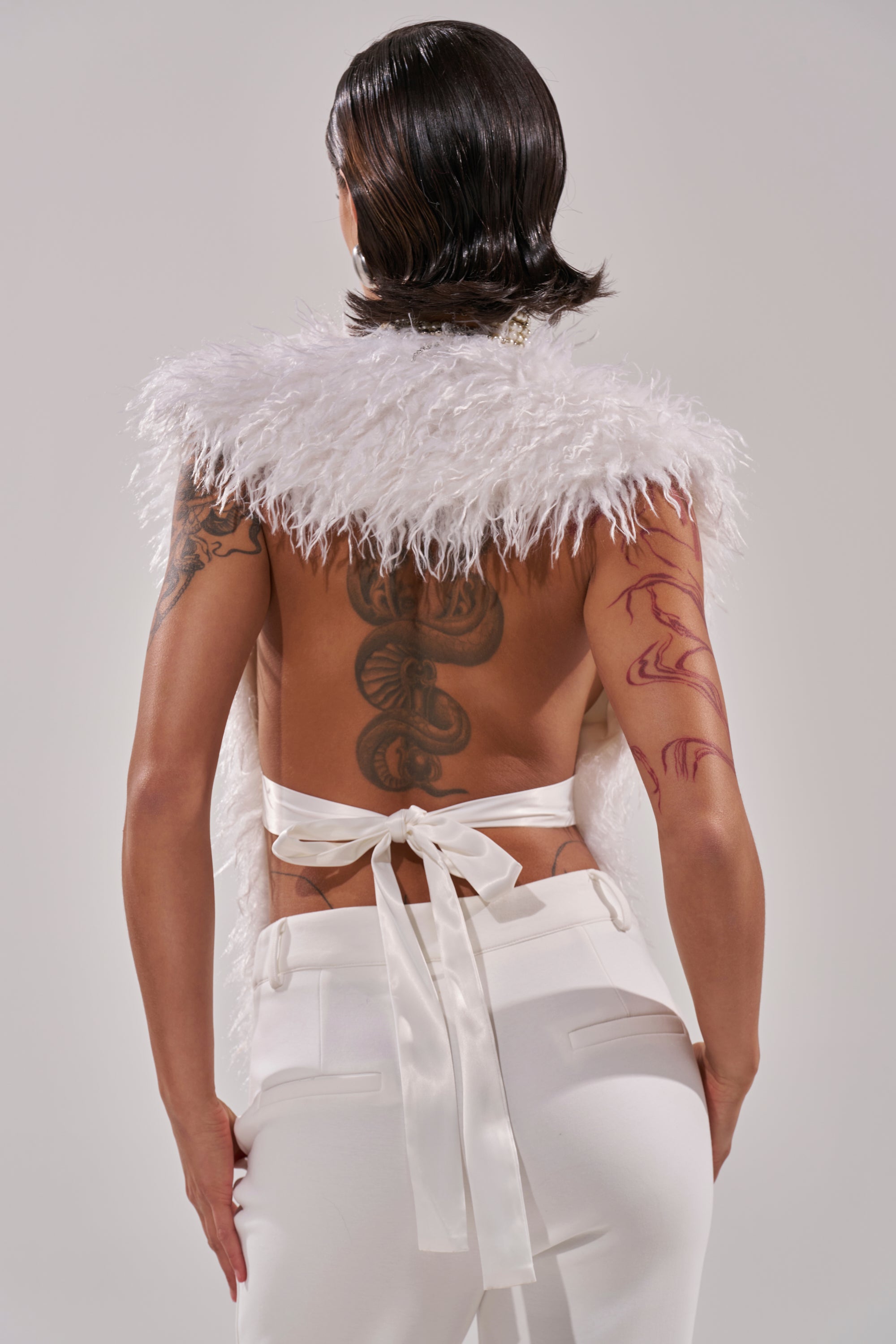 A person faces away in white pants and the JAMEELA FAUX FUR TOP, which is ivory with an open back tied by a satin ribbon. Back and left arm tattoos are visible. The background is plain light gray.