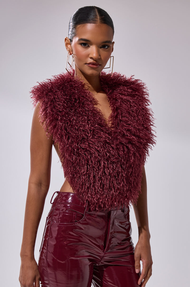 A woman confidently models the TARA FAUX FUR HALTER TOP in deep burgundy, styled with matching glossy pants, large geometric earrings, and slicked-back hair against a simple light background.
