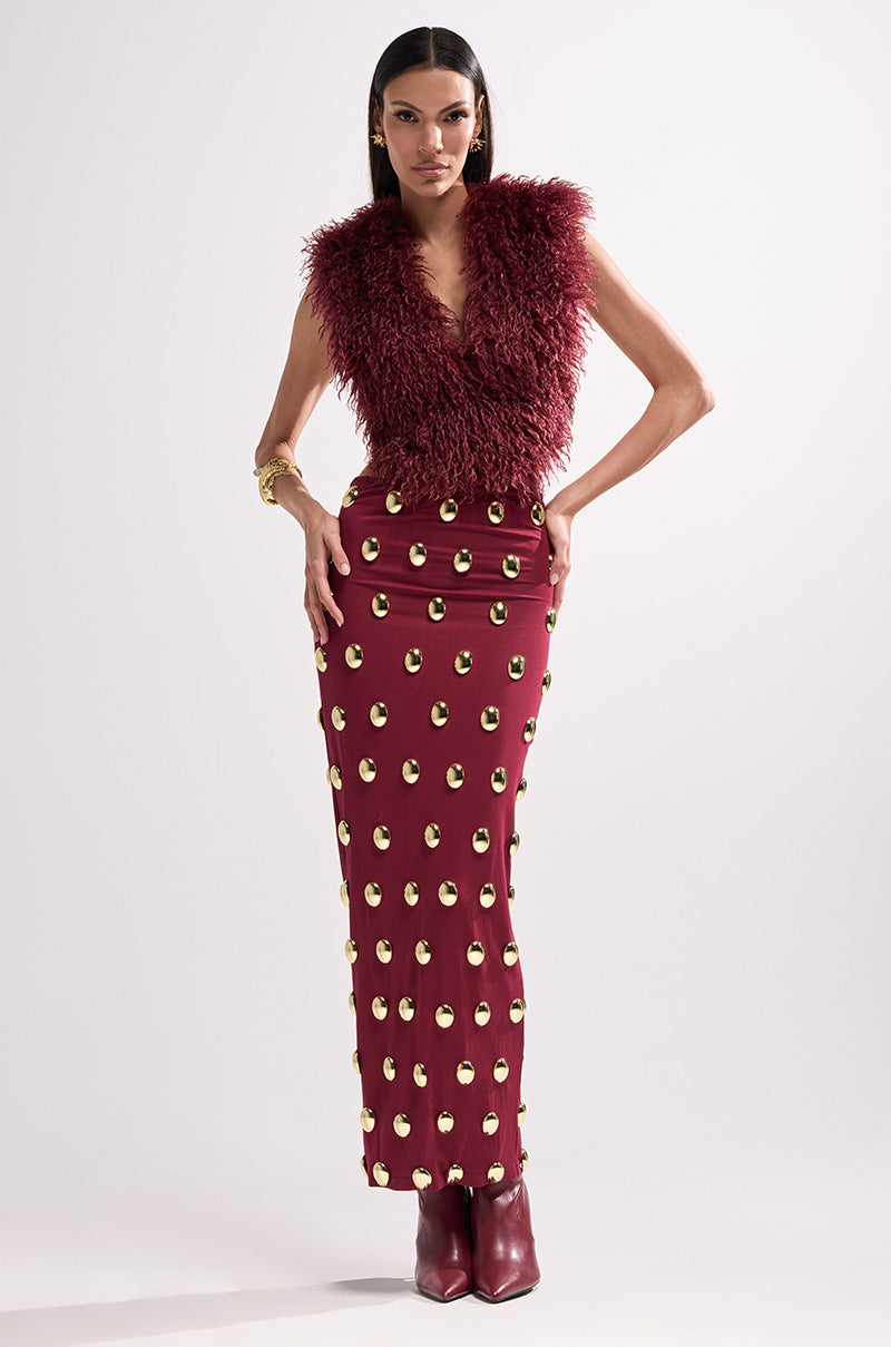 A woman poses confidently in the Tara Faux Fur Halter Top, a wine red fitted piece paired with a skirt detailed with gold circles. She completes her look with burgundy ankle boots and gold jewelry against a plain background.