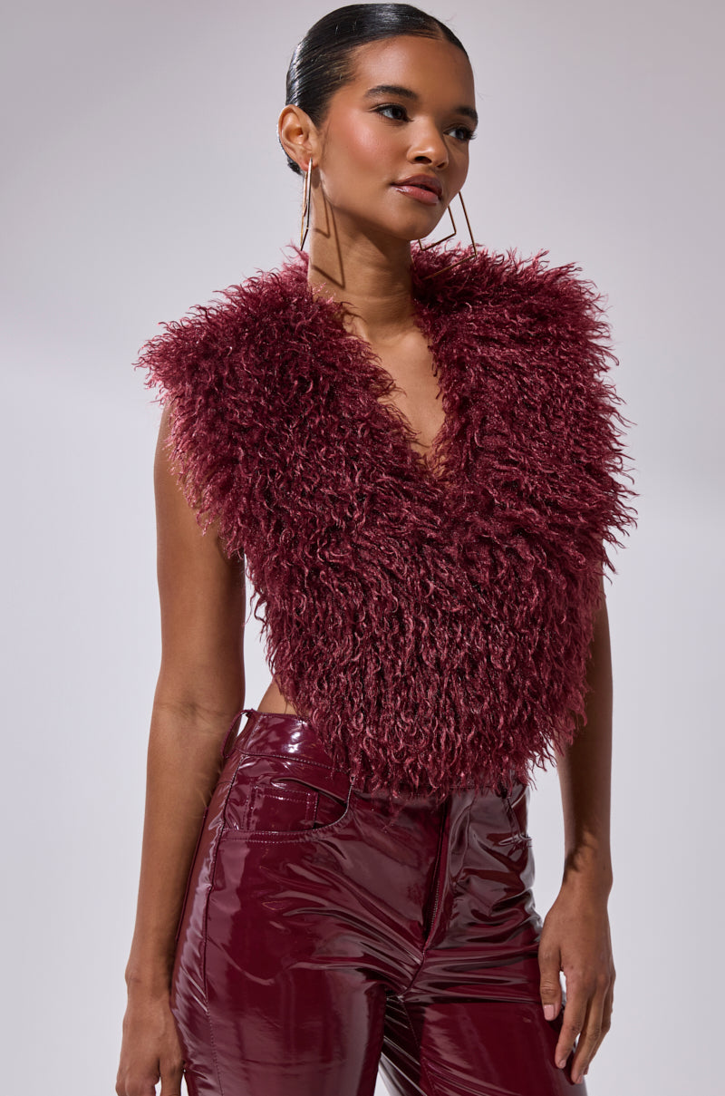 A woman wears the TARA FAUX FUR HALTER TOP in wine-red, styled with glossy burgundy pants, slicked-back hair, and large hoop earrings against a plain light background.