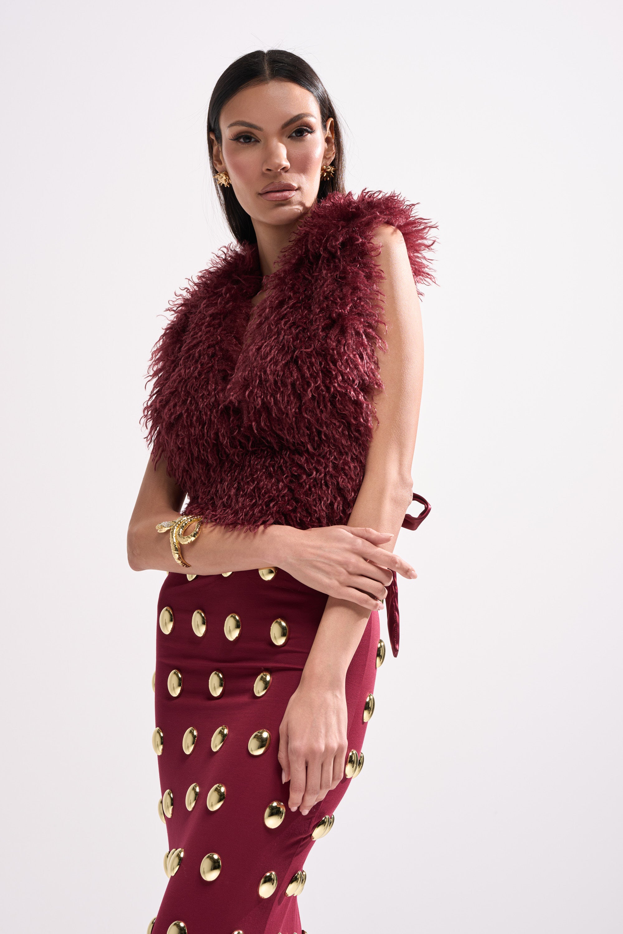 A woman wears the TARA FAUX FUR HALTER TOP in wine-red with a matching skirt featuring large gold buttons, gold jewelry, and a satin ribbon tie, posing against a plain light background with straight dark hair.