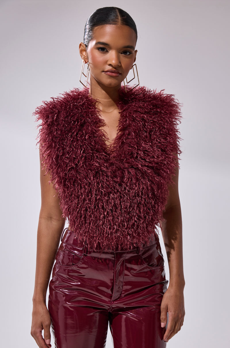 A woman wears the TARA FAUX FUR HALTER TOP in wine red with a satin ribbon tie, paired with shiny burgundy vinyl pants. She poses against a plain light background, sporting sleek hair, hoop earrings, and a confident expression.