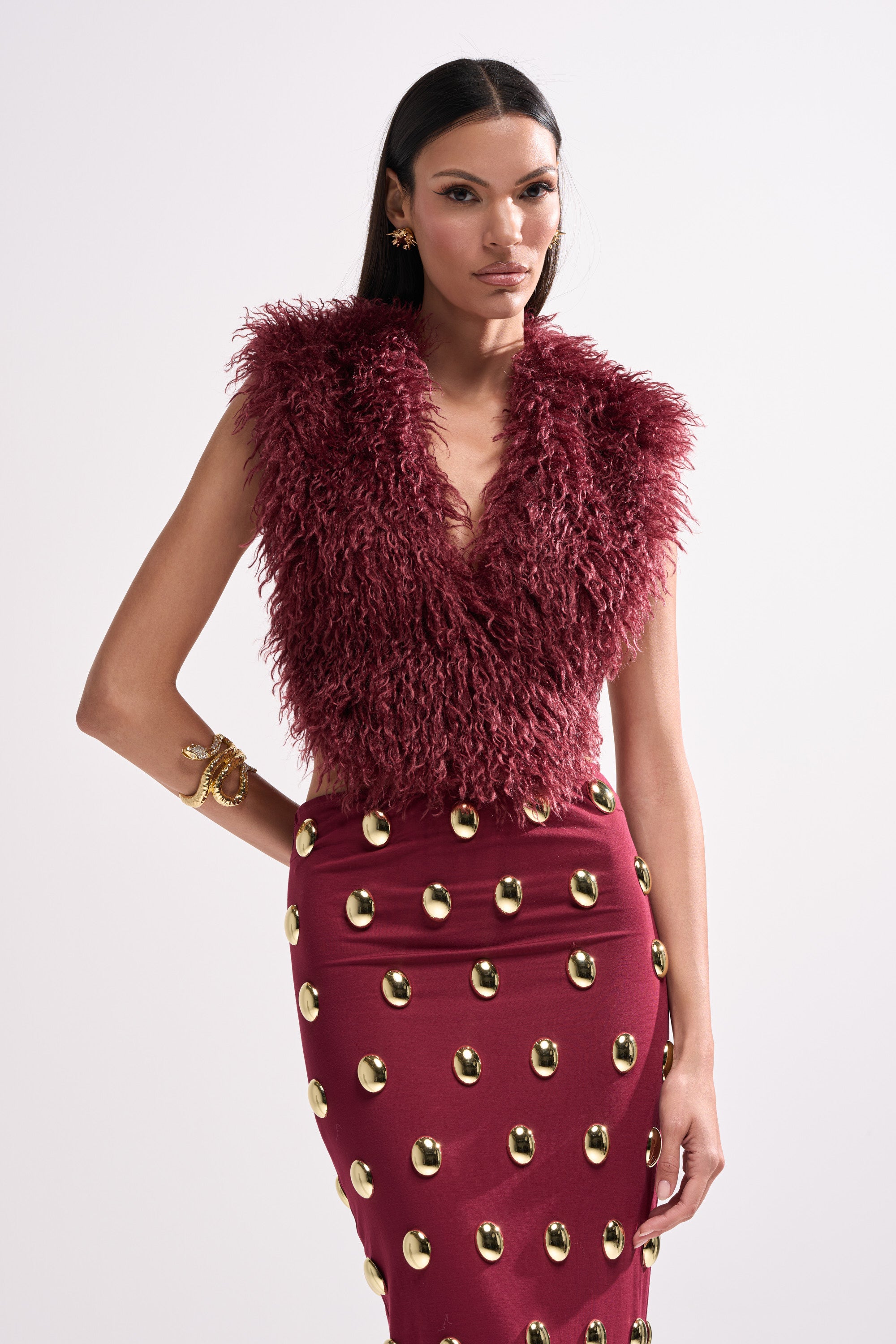 Wearing the TARA FAUX FUR HALTER TOP in wine-red with a satin ribbon tie and a matching high-waisted skirt featuring gold oval embellishments, a woman poses against a plain white background.