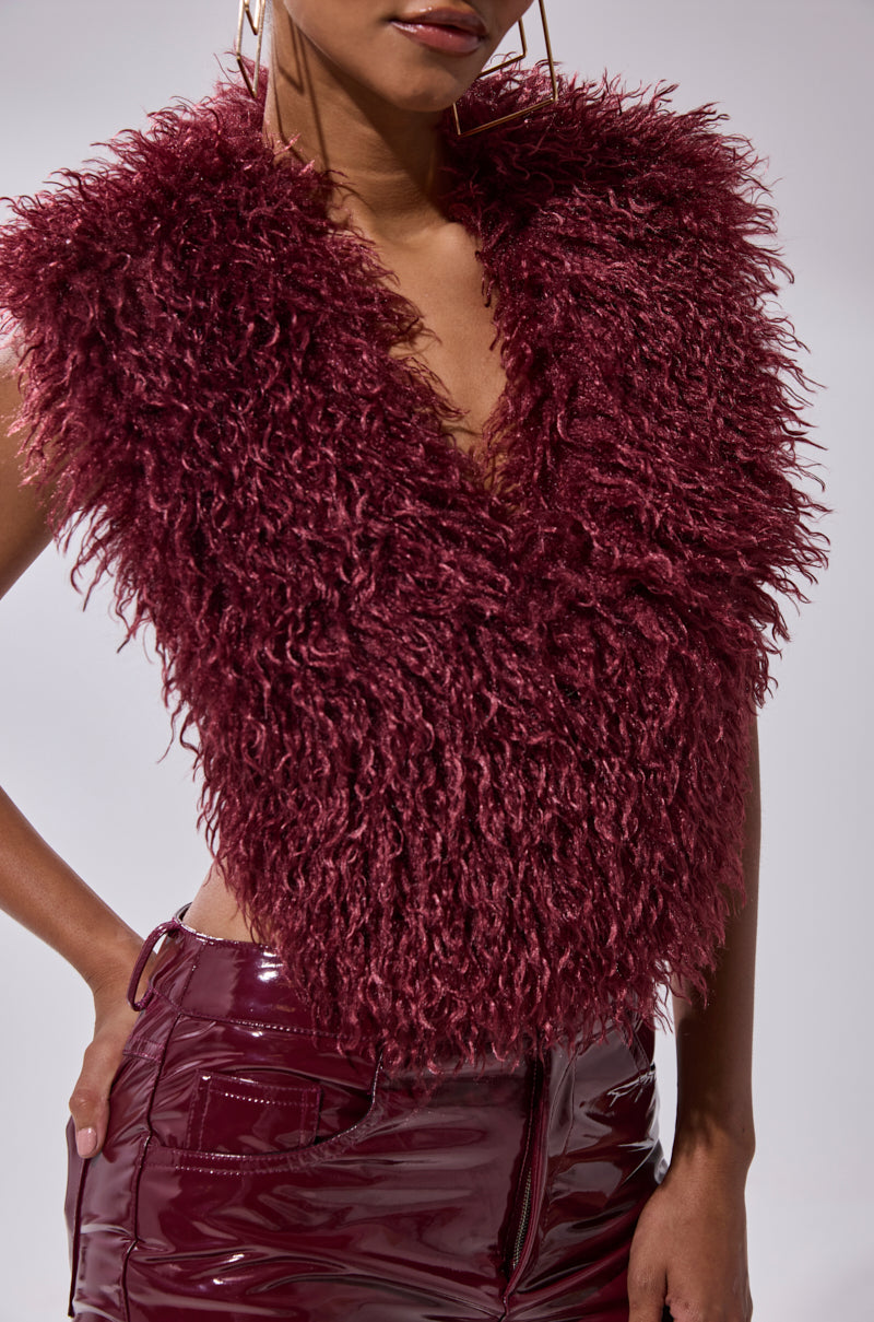 Wearing the TARA FAUX FUR HALTER TOP in wine red with shiny, high-waisted burgundy vinyl pants, a person stands against a neutral background, hand on hip and face partially out of frame.
