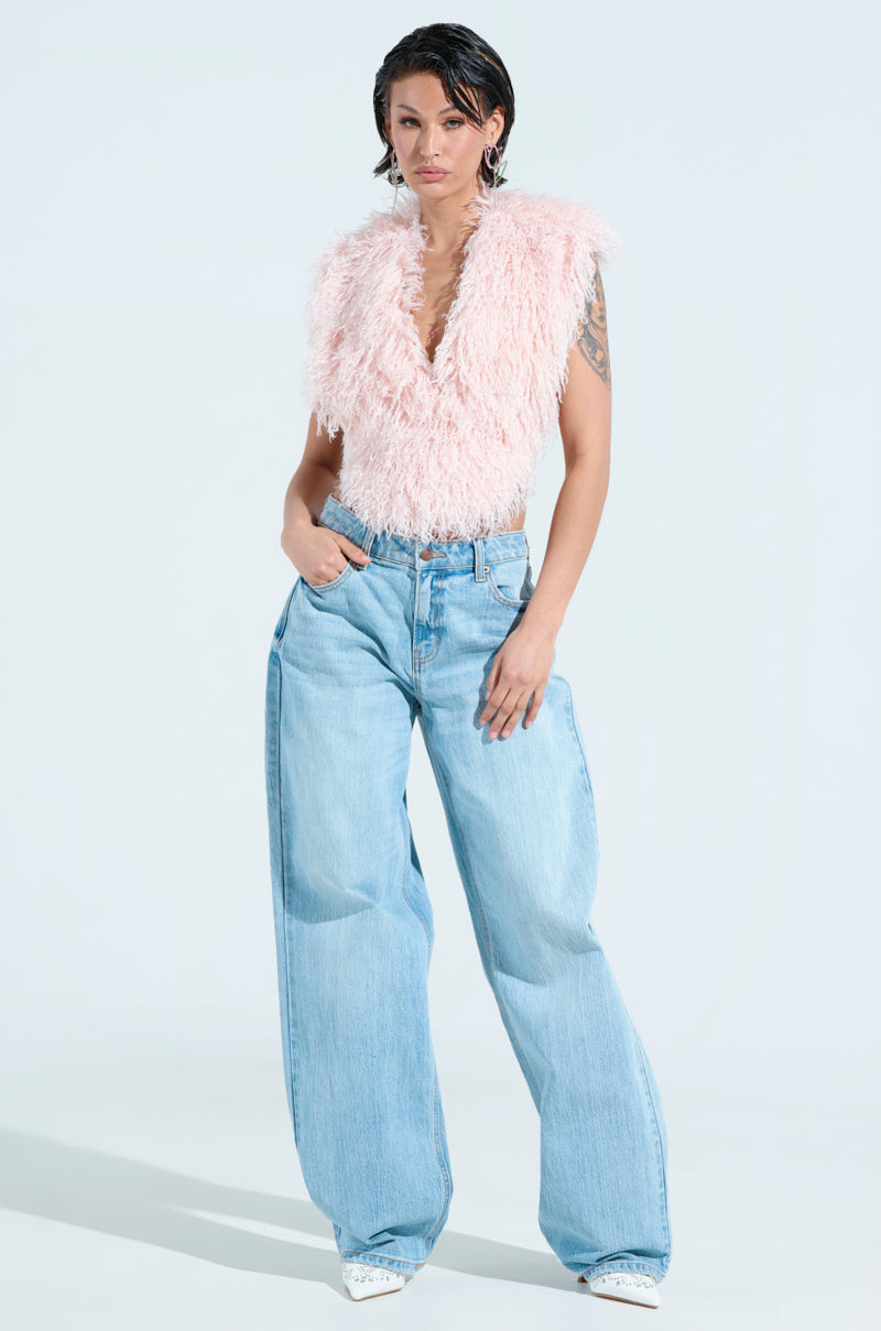 A person poses against a light background, wearing the pink JAMEELA TOP featuring a plunging V-neckline, paired with light blue wide-leg jeans and white shoes. They have short dark hair and stand with one hand in their pocket. 9