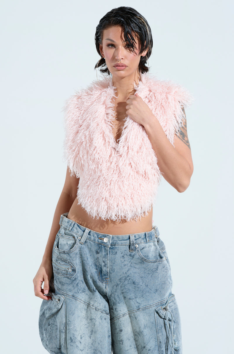 A person with short, dark wet hair poses confidently in the JAMEELA TOP, a fluffy, sleeveless pink faux fur top, paired with baggy acid-wash jeans featuring large pockets against a plain white background. 1
