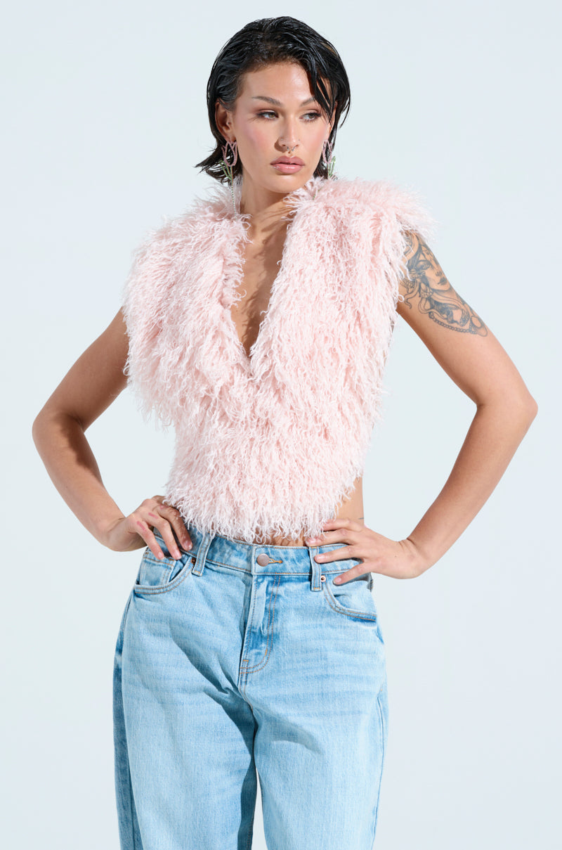 A person confidently poses with hands on hips, wearing the JAMEELA TOP, a fluffy pink faux fur halter top, paired with light blue jeans. They sport wet hair, dangling earrings, and an arm tattoo against a plain light background. 2