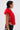 A person with short, wet-look dark hair and large hoop earrings wears the bright red CARLA FAUX FUR HALTER TOP in a cropped, sleeveless style with black jeans featuring a graphic patch, standing in profile against a plain background.