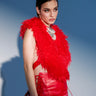 A woman with dark hair wears the bold, cropped CARLA FAUX FUR HALTER TOP in red and matching high-waisted red leather pants with lace-up sides, accessorized with a chunky metallic necklace, against a blue background.