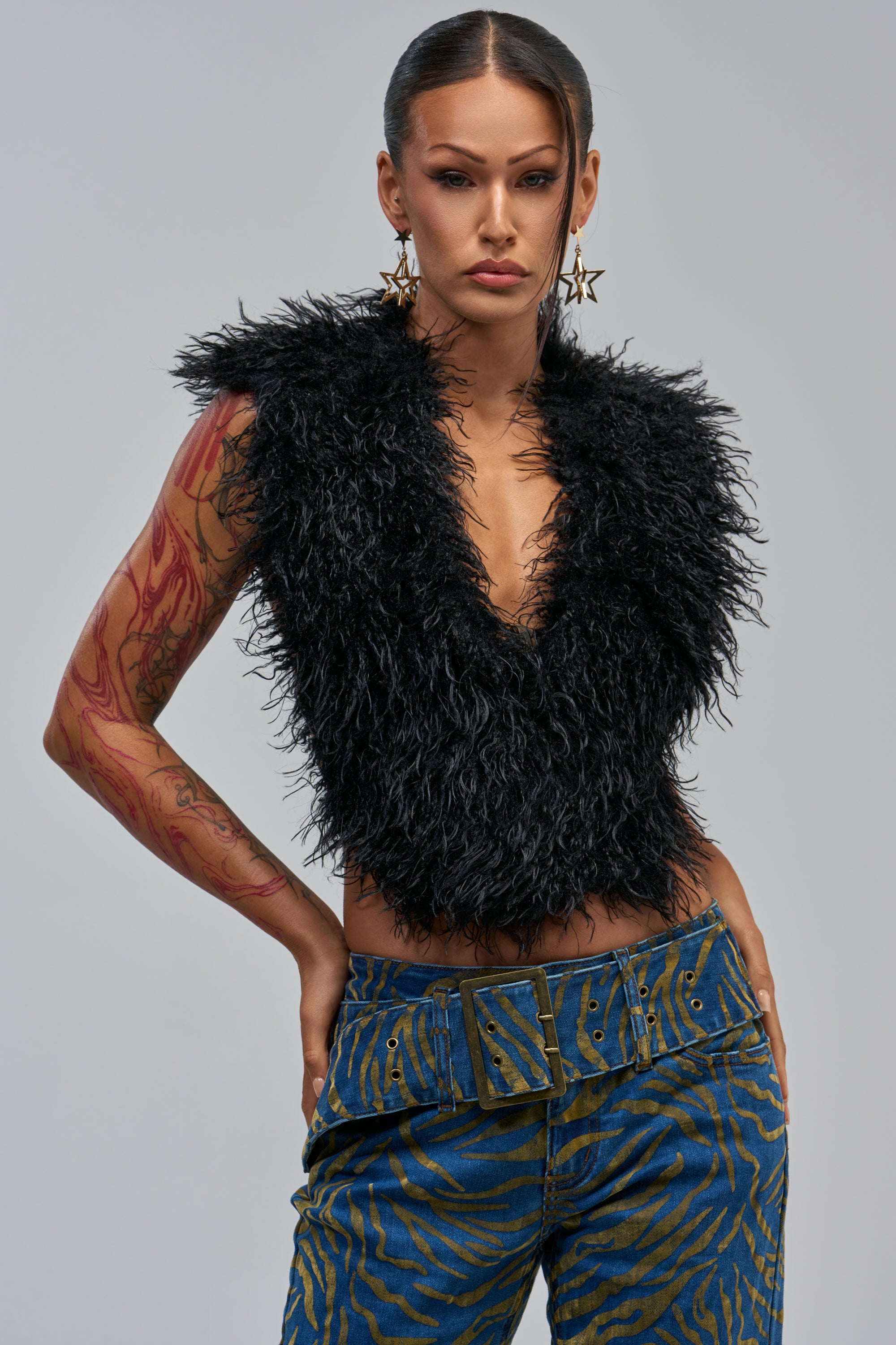 Woman with slicked-back hair wears star earrings, the TEMPLETON TOP in black faux fur, blue patterned pants with a wide belt, and shows red tattoos on her left arm against a plain gray background.