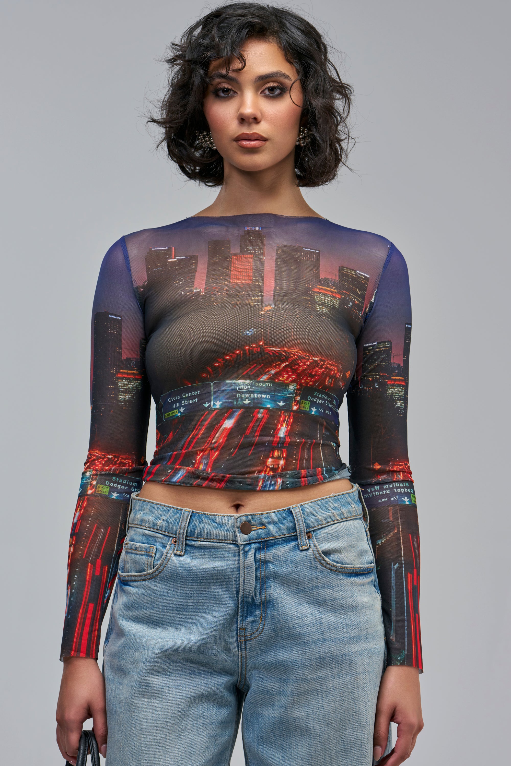 A person wears the AT MY OWN PACE MESH LONG SLEEVE TOP, a long-sleeved, sheer mesh garment featuring a nocturnal city skyline and highway traffic print.