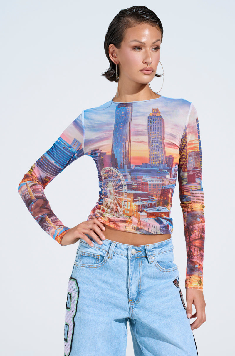 Wearing the CITY LIGHTS MESH LONG SLEEVE TOP with city graphics and light blue jeans, a short-haired person stands against a clear sky. They sport large hoop earrings and look to the side with one hand on their hip.