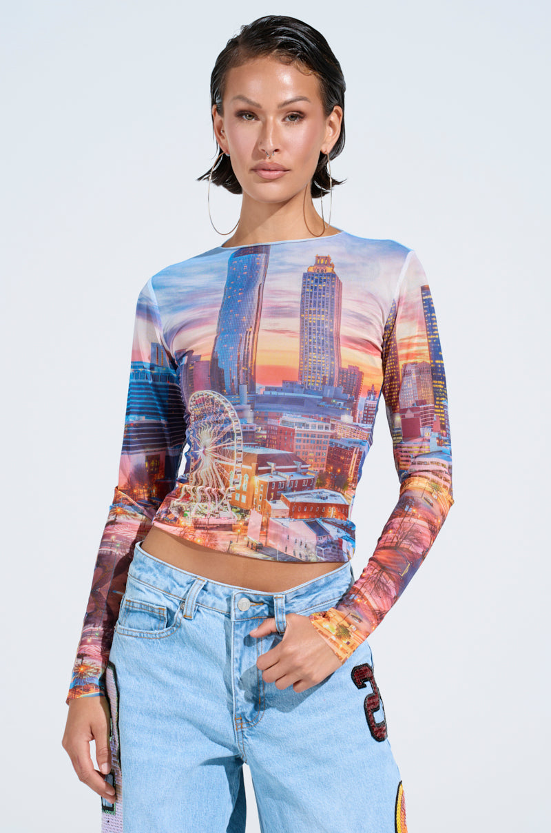 A person with short dark hair wears the CITY LIGHTS MESH LONG SLEEVE TOP, showcasing a vibrant cityscape print, paired with light blue patched jeans. The stretchy fabric ensures comfort, while a plain background accentuates the striking outfit.