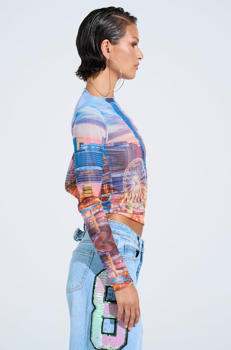 Side profile of a person with short, slicked-back hair and hoop earrings, wearing a CITY LIGHTS MESH LONG SLEEVE TOP with a cityscape design and light blue jeans featuring an embellished number 8.