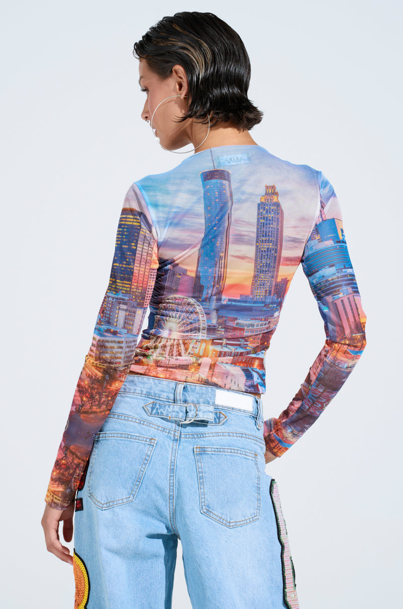 A person with short hair is wearing the fitted, stretchy CITY LIGHTS MESH LONG SLEEVE TOP and light blue jeans with graphic patches. They have hoop earrings and face away from the camera, highlighting the top's colorful urban city design.