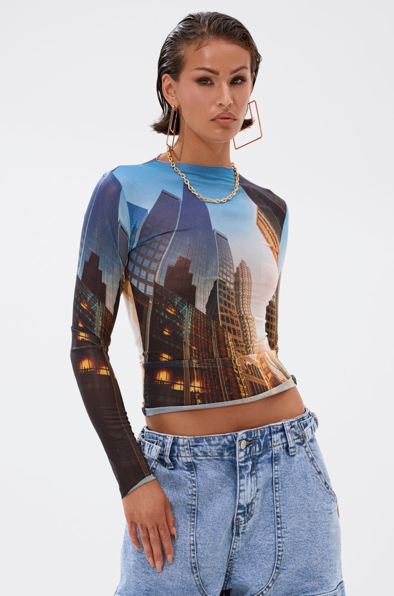 A woman with slicked-back hair models the BIG CITY LONG SLEEVE MESH TOP with a city graphic, paired with high-waisted loose blue jeans, large square hoop earrings, and a gold chain necklace against a plain background. 0