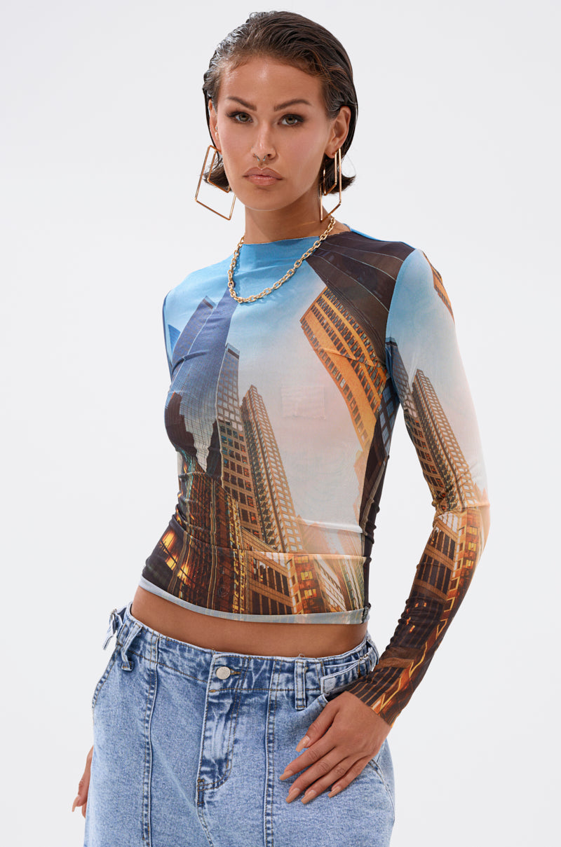 A woman wearing the BIG CITY LONG SLEEVE MESH TOP, light denim jeans, large gold hoop earrings, and a gold chain necklace stands against a plain light background. 1