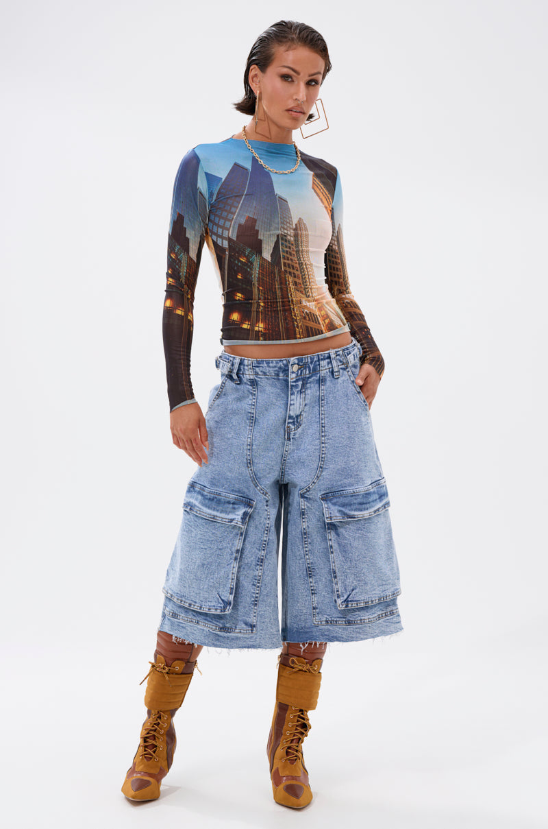 A woman stands confidently in the BIG CITY LONG SLEEVE MESH TOP, paired with baggy knee-length denim cargo shorts, brown lace-up boots, and large geometric earrings against a plain white background. 5
