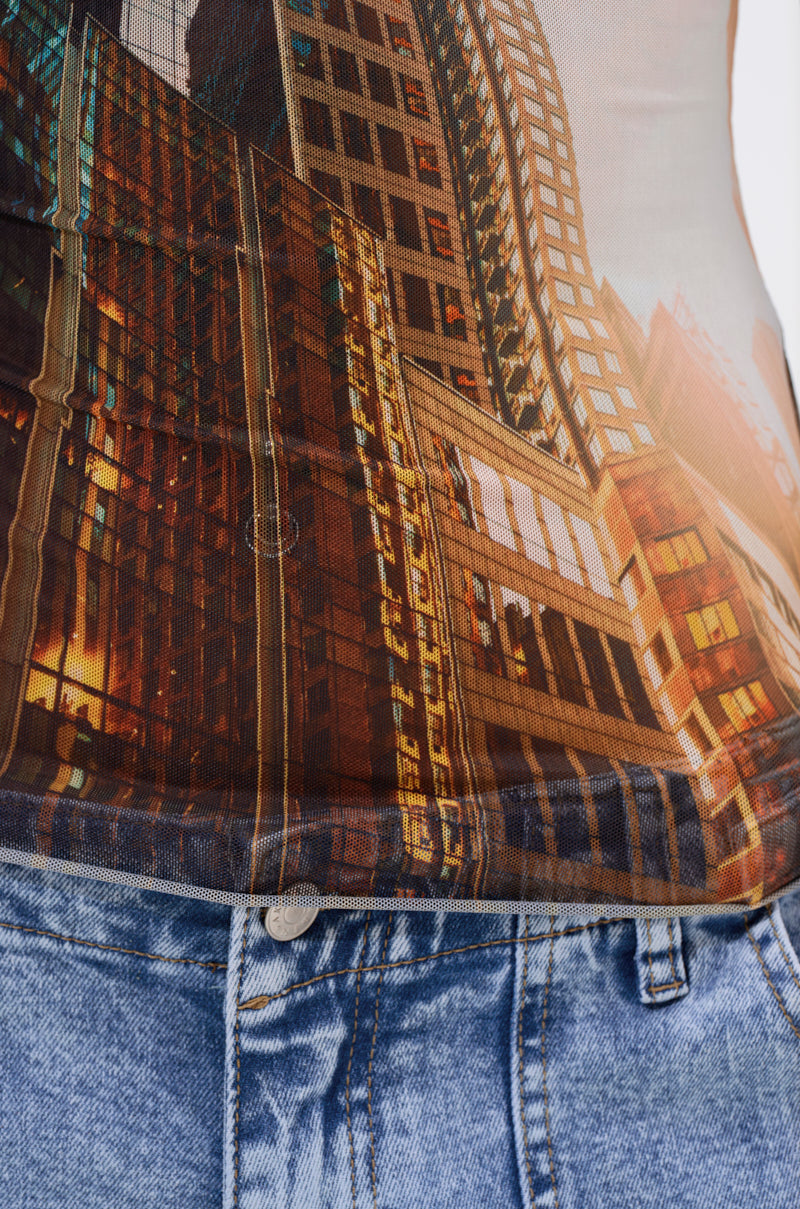 Close-up of a person wearing the BIG CITY LONG SLEEVE MESH TOP featuring a cityscape print with tall buildings illuminated by warm lights for an urban graphic effect, paired with blue jeans. 8