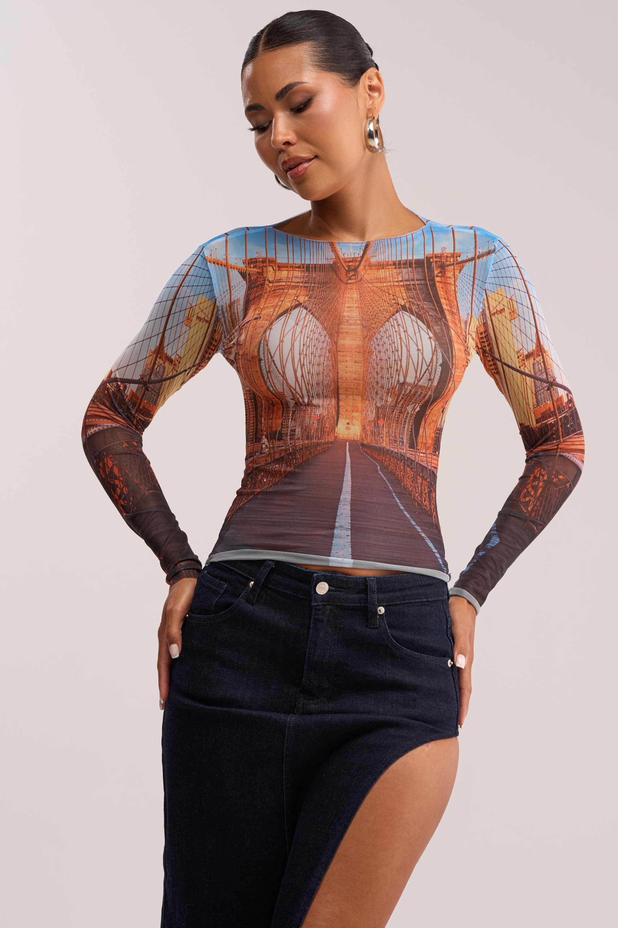 A woman models the ROADTRIP MESH LONG SLEEVE TOP with a Brooklyn Bridge graphic, styled for a summer date night with a high-slit dark denim skirt, hoop earrings, and slicked-back hair against a plain background.