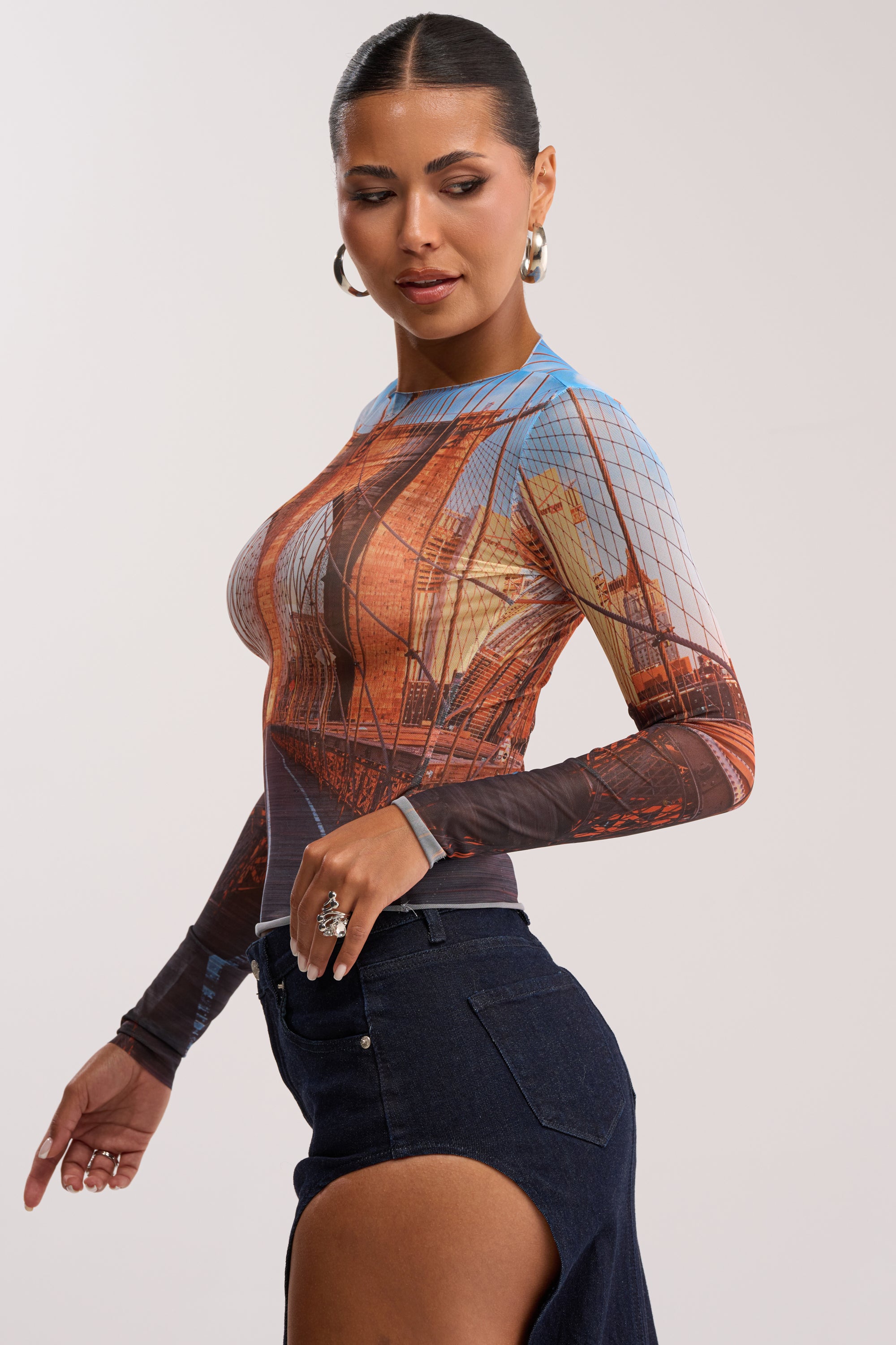 A woman with slicked-back hair wears hoops, the ROADTRIP MESH LONG SLEEVE TOP—ideal for a summer date night—and a dark denim skirt with a high slit, posing sideways against a plain light background.