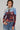 A person strikes a pose against a plain backdrop, showcasing urban-chic fashion in the CITYSCAPE TOP, featuring a Chicago skyline graphic. They enhance their style with a chunky chain necklace and oversized hoop earrings. Their short hair perfectly complements the ensemble as they confidently stand with one hand in the pocket of their faded jeans.