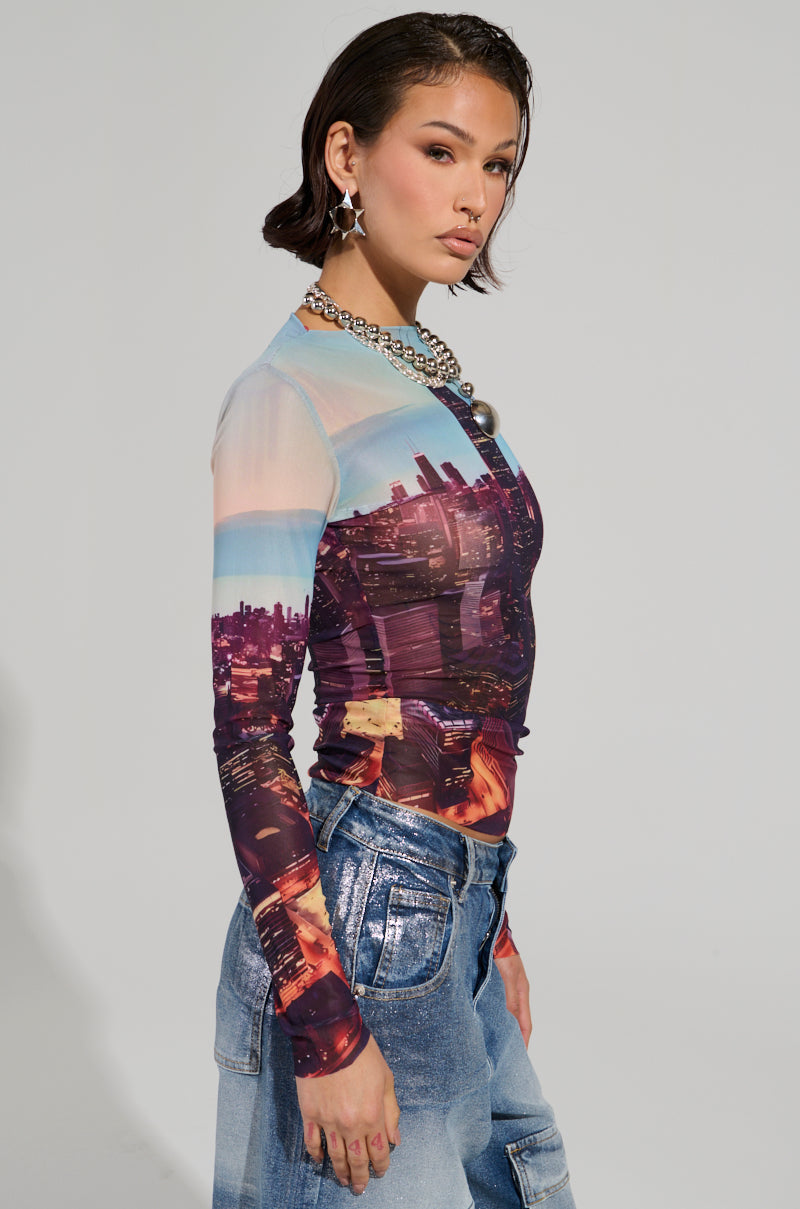 A person with short dark hair, wearing a long-sleeved CITYSCAPE TOP showcasing a Chicago skyline graphic and denim jeans, poses against a plain background. They accessorize with hoop earrings and a chunky necklace, exuding urban-chic fashion while looking confidently at the camera.