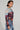 A person with short dark hair, wearing a long-sleeved CITYSCAPE TOP showcasing a Chicago skyline graphic and denim jeans, poses against a plain background. They accessorize with hoop earrings and a chunky necklace, exuding urban-chic fashion while looking confidently at the camera.