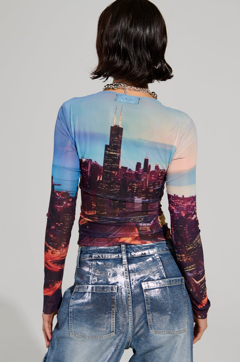 A person with short, dark hair is facing away, wearing the long-sleeve CITYSCAPE TOP featuring a Chicago skyline graphic. They also wear high-waisted, shiny textured, faded blue jeans that embody the essence of urban-chic fashion.
