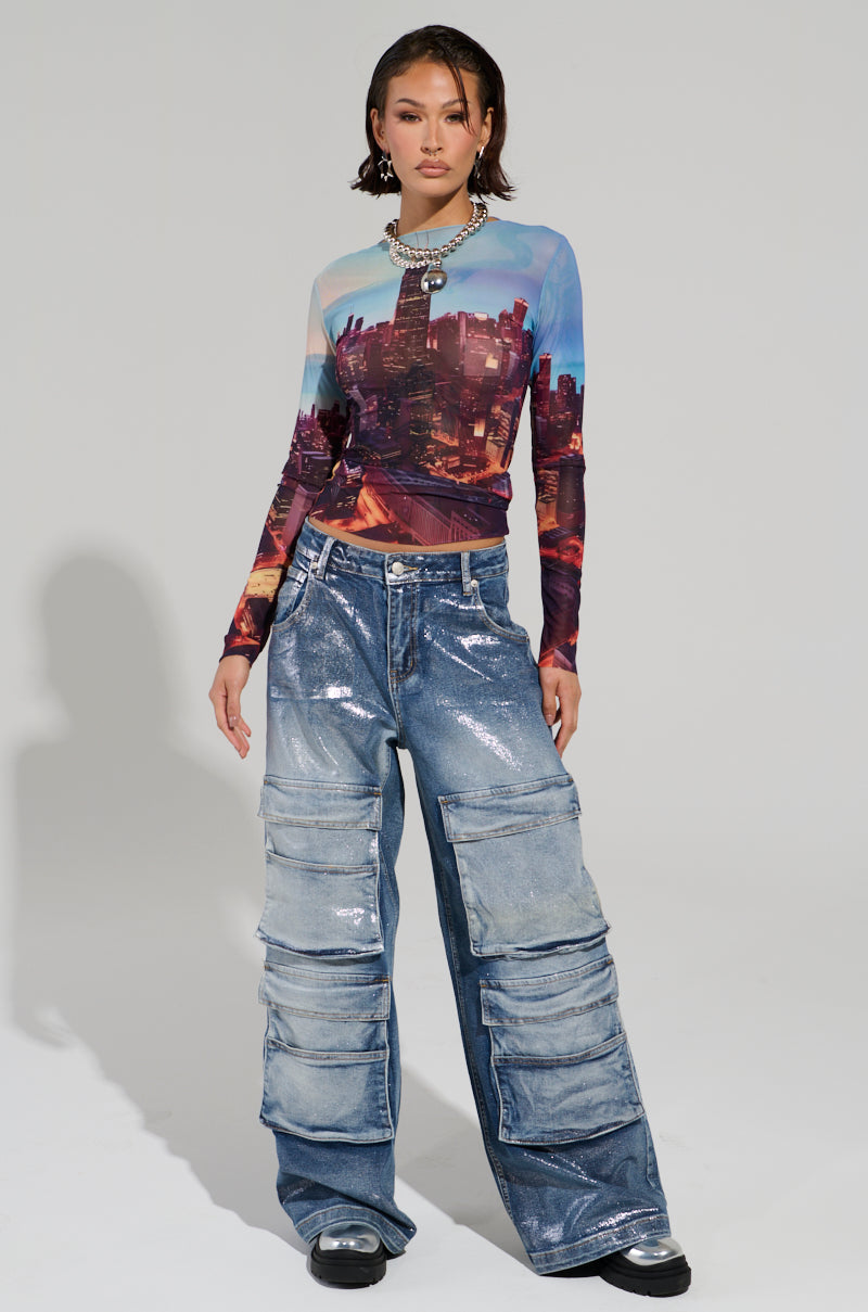 A person stands in a studio wearing the CITYSCAPE TOP, featuring a Chicago skyline graphic on a sheer mesh fabric, paired with shiny, oversized cargo jeans. They have short hair and large hoop earrings. The plain background highlights the urban-chic fashion of the outfit.