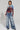 A person stands in a studio wearing the CITYSCAPE TOP, featuring a Chicago skyline graphic on a sheer mesh fabric, paired with shiny, oversized cargo jeans. They have short hair and large hoop earrings. The plain background highlights the urban-chic fashion of the outfit.