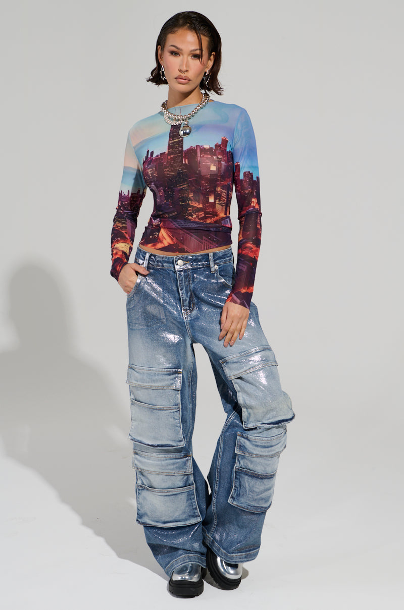A person exudes confidence in an urban-chic fashion ensemble, wearing the CITYSCAPE TOP—a long-sleeve shirt adorned with a Chicago skyline graphic—and oversized blue jeans featuring multiple pockets. They complete the look with a statement necklace and black shoes, set against a plain backdrop.