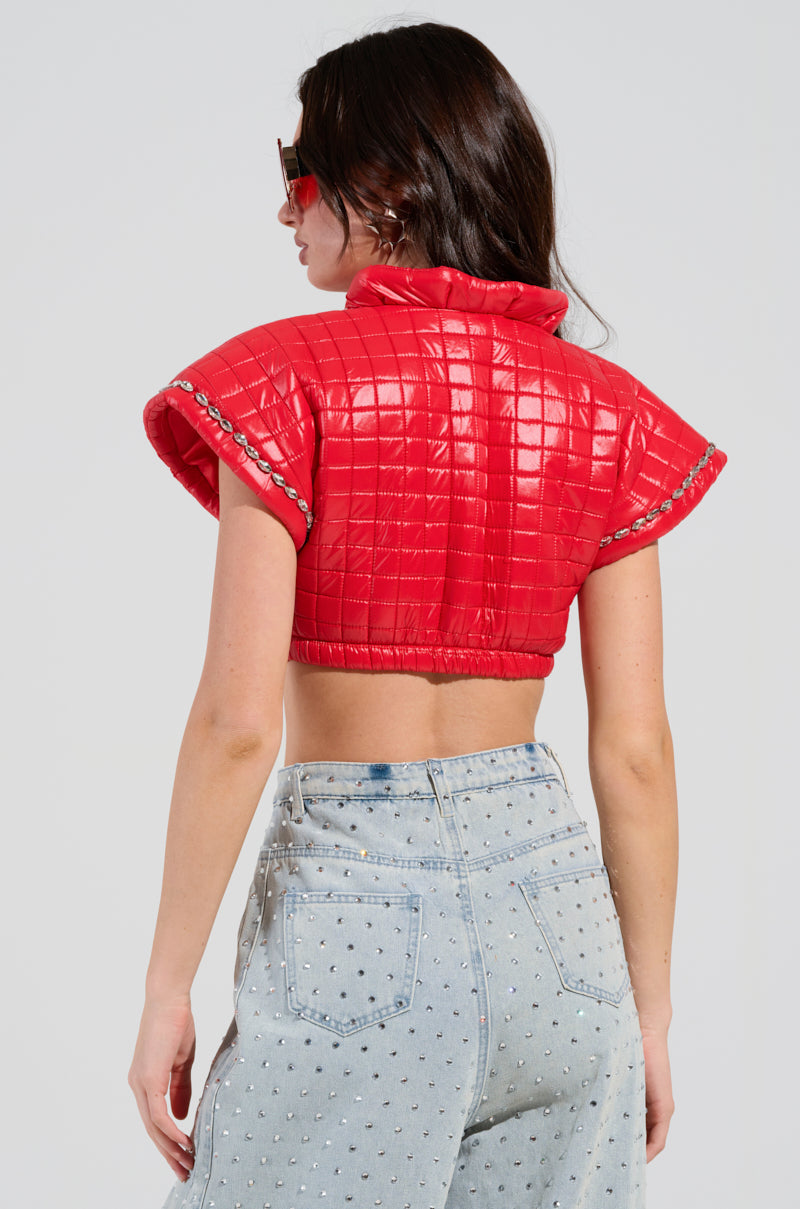 A person with long hair is wearing the PROTECT YOUR ENERGY STATEMENT BLOUSE, a red, quilted cropped silhouette adorned with chain details around the sleeves and a rhinestone trim. They have paired it with light blue, high-waisted jeans featuring small embellishments and are donning stylish sunglasses against a plain backdrop.