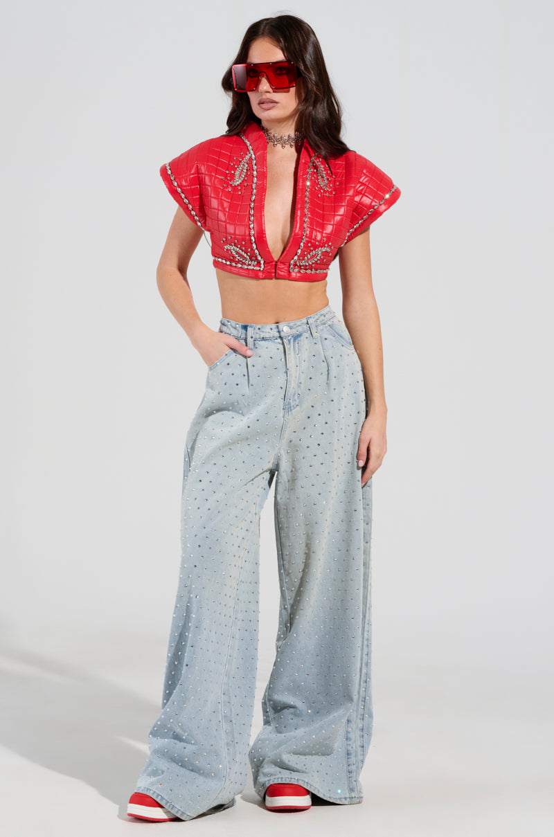 A woman models a vibrant, patterned AKIRA Label PROTECT YOUR ENERGY STATEMENT BLOUSE with short sleeves, paired with light blue, wide-legged pants embellished with rhinestone trim. She accessorizes her look with red sunglasses and matching red sneakers.