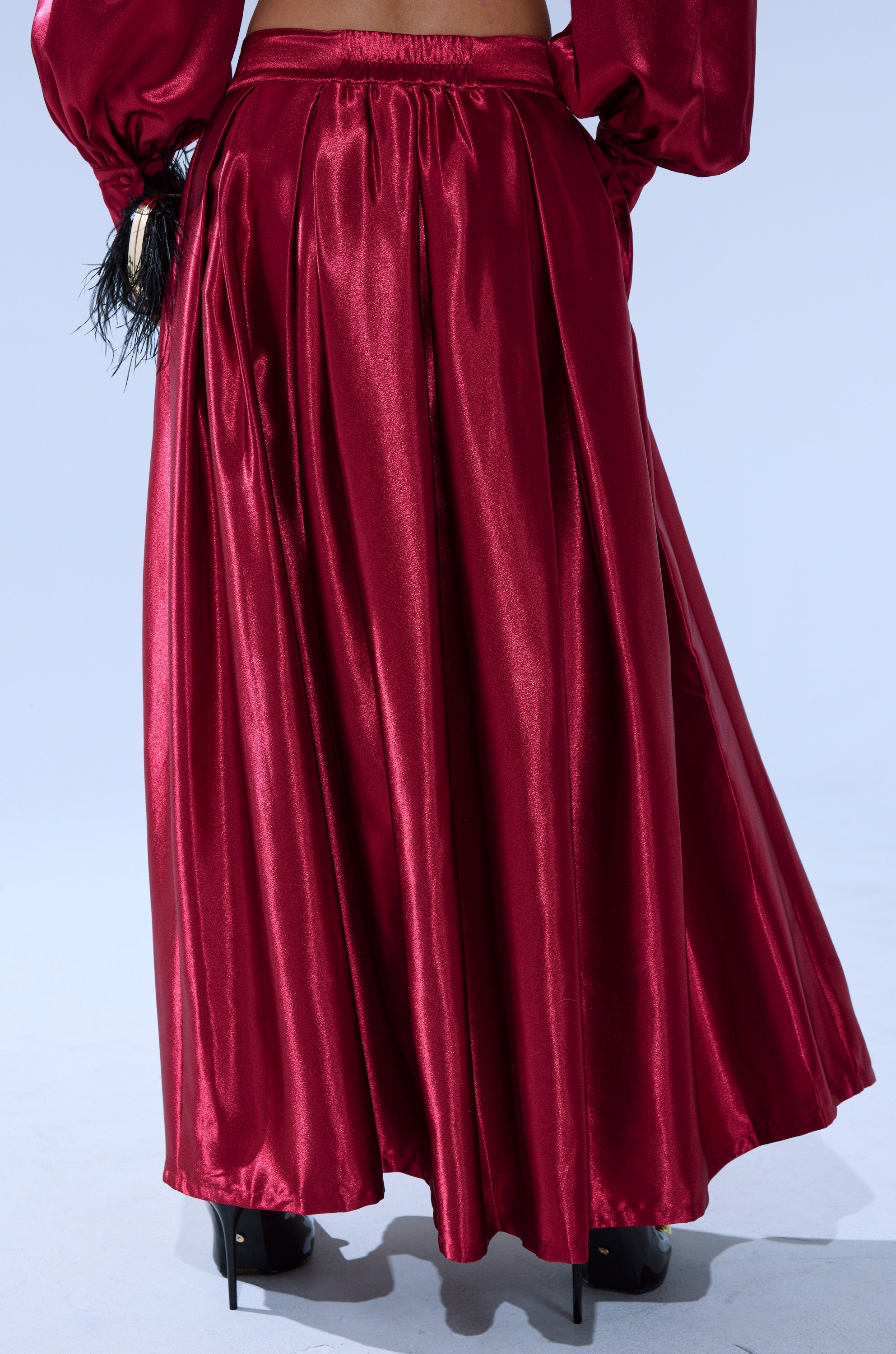 A person wears the CANDICE SATIN MAXI SKIRT, a long, shiny red skirt with gathered fabric and a fitted waistband, paired with high heels and a matching red blouse with voluminous sleeves for an elegant look. 1