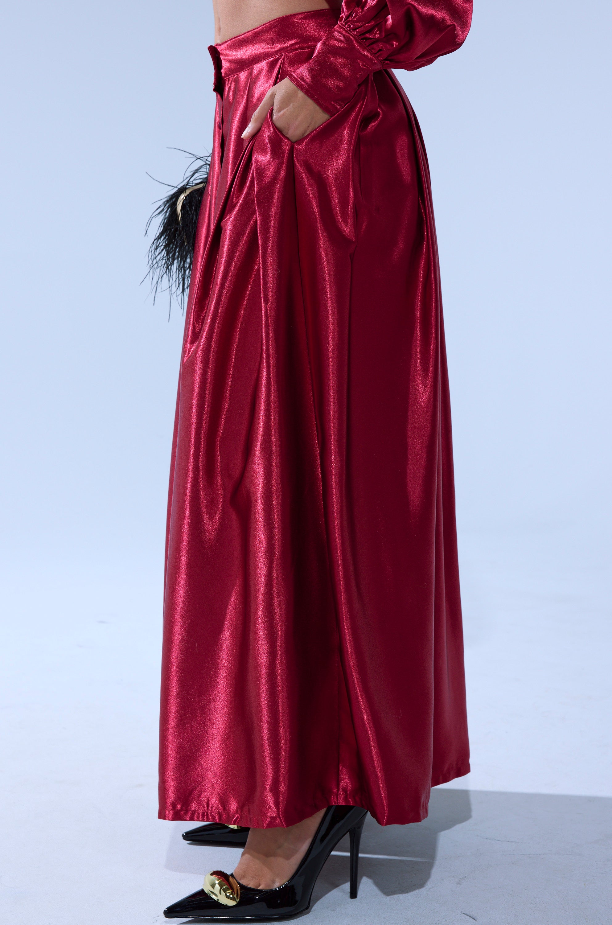 Wearing the CANDICE SATIN MAXI SKIRT in shiny red with pleats, a hand in the pocket, black pointed gold-detailed heels, and holding a small black feathered clutch against a plain light gray background. 2