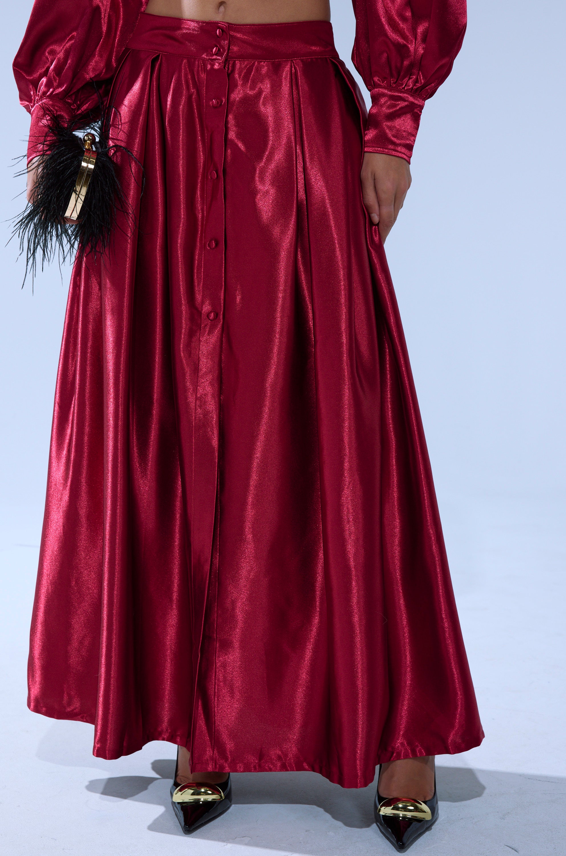 A person in the CANDICE SATIN MAXI SKIRT—a shiny red satin maxi with front buttons—holds a black feathered clutch and wears gold pointed-toe heels. 3