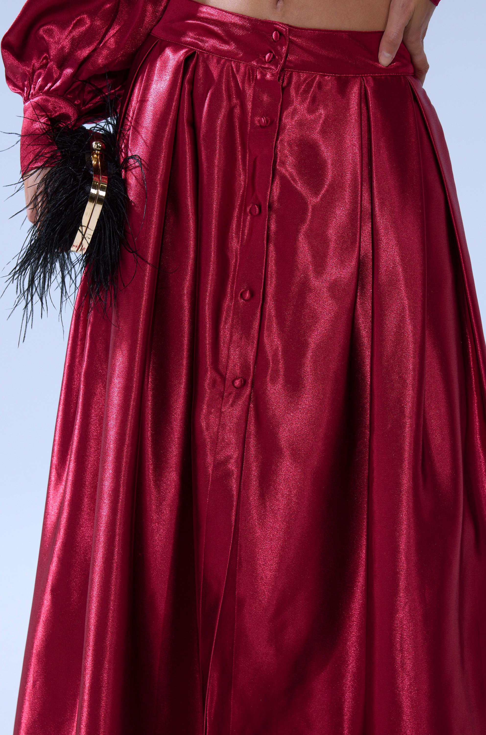 A close-up of a person wearing the CANDICE SATIN MAXI SKIRT, a shiny red high-waisted skirt with front buttons. Part of a matching top and a hand holding a black feathered clutch are also visible. 6