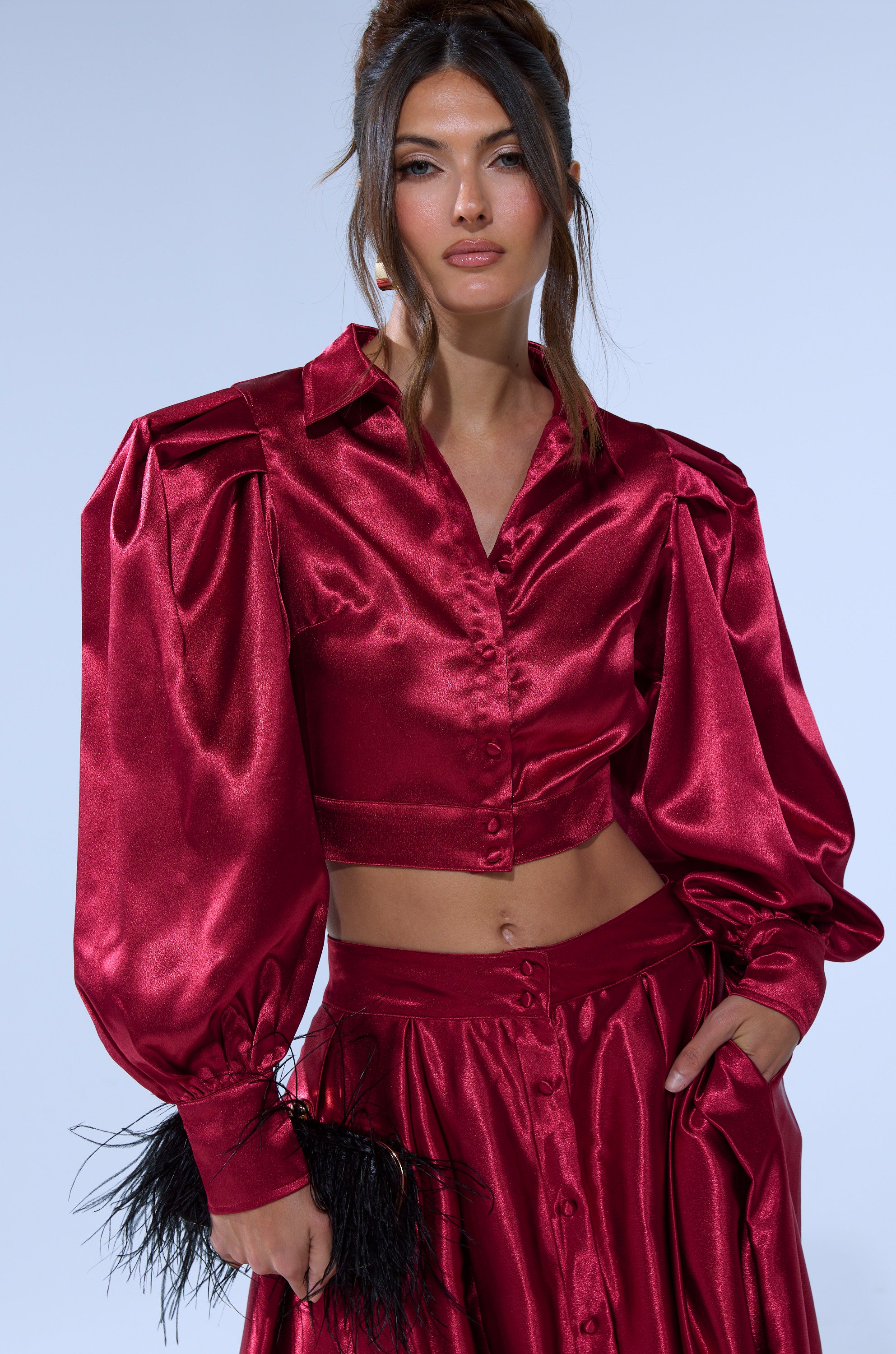 A woman wears the CANDICE OPEN BACK SATIN BUTTON DOWN SHIRT in shiny deep red with a matching high-waisted skirt, holding a black feathered accessory. Her long brown hair is styled half-up, half-down.