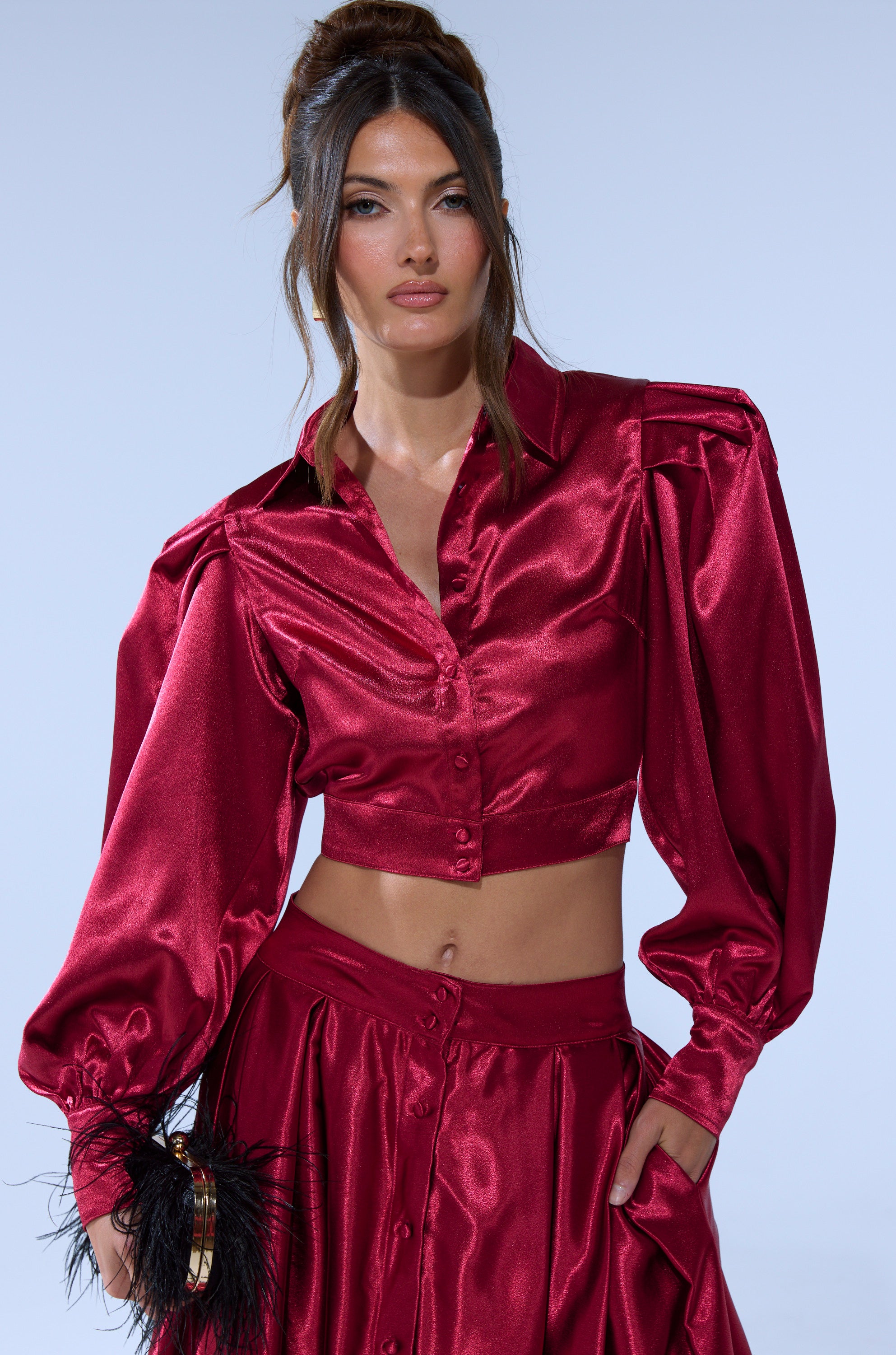 A woman stands against a light blue background wearing the CANDICE OPEN BACK SATIN BUTTON DOWN SHIRT in shiny deep red, paired with a matching high-waisted skirt and holding a small purse with black feather accents.