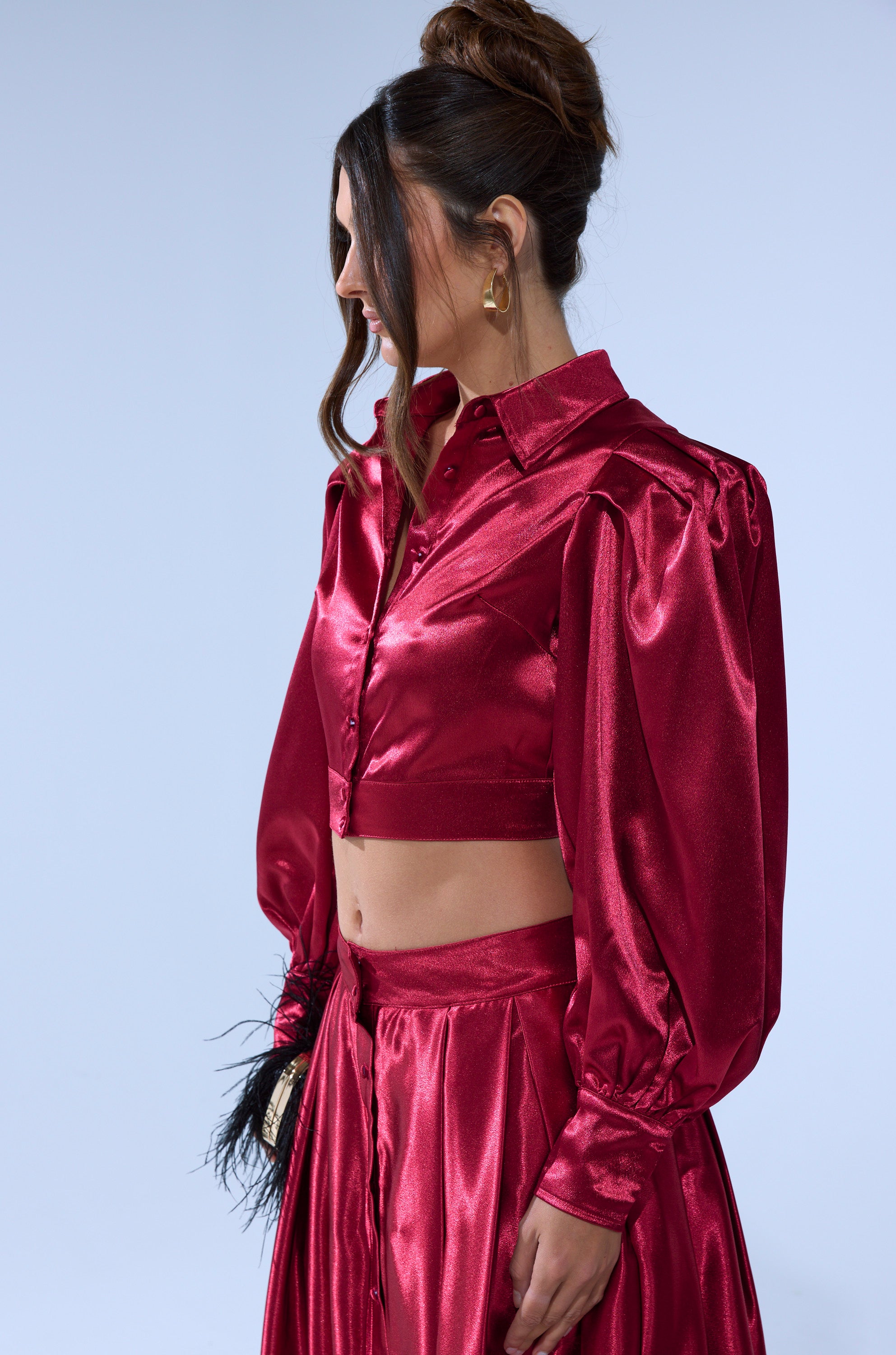 A woman with long brown hair in an updo wears a shiny red Candice Satin Maxi Skirt and a matching Candice Open Back Satin Button Down Shirt, facing sideways and looking away from the camera.
