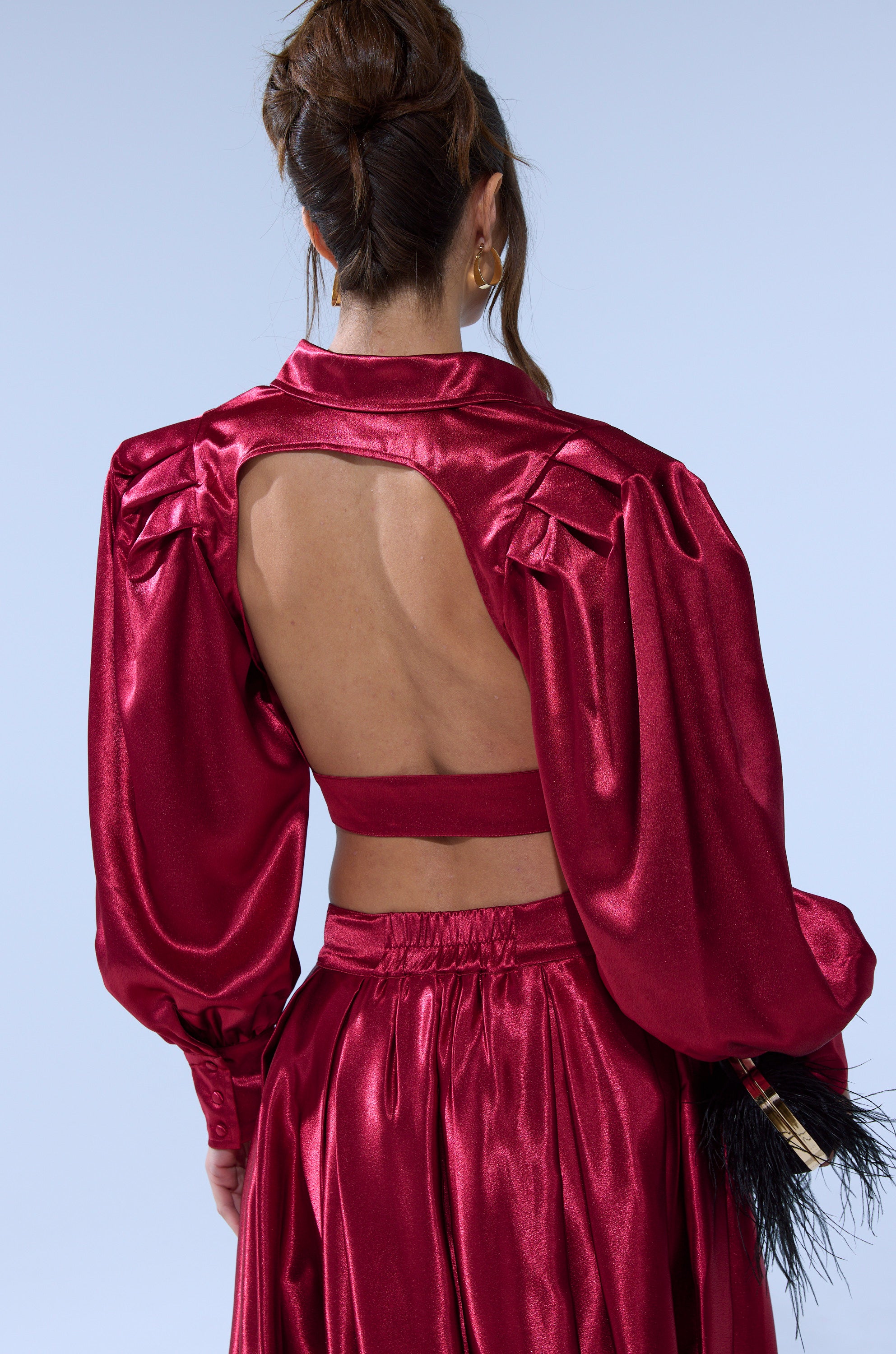 A woman with brown hair in an updo wears the CANDICE OPEN BACK SATIN BUTTON DOWN SHIRT, a shiny deep red cropped top with voluminous sleeves and an open back, paired with a matching satin-look skirt.