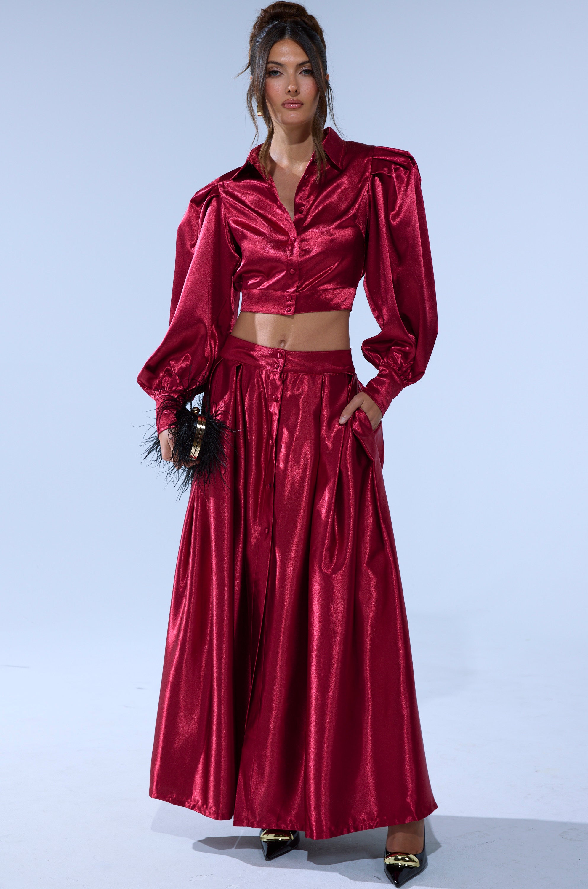 A woman poses against a plain background in the CANDICE OPEN BACK SATIN BUTTON DOWN SHIRT, styled in deep red with long sleeves, paired with a matching skirt, black pointed heels, and a small feathered clutch.