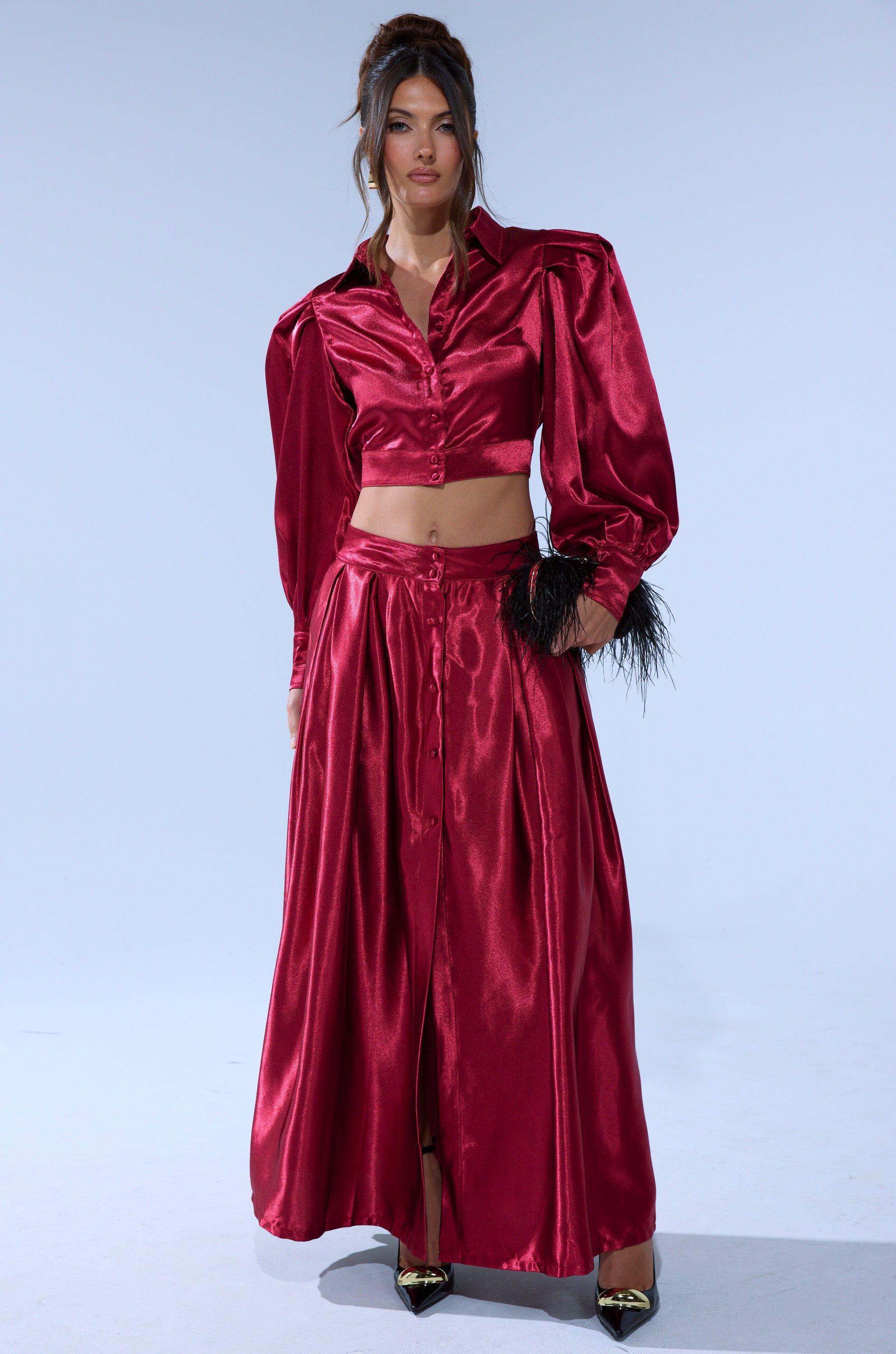 A woman wears the CANDICE OPEN BACK SATIN BUTTON DOWN SHIRT, a shiny red top with puff sleeves and black feather accents on one sleeve, paired with a long skirt. She stands against a plain light background.
