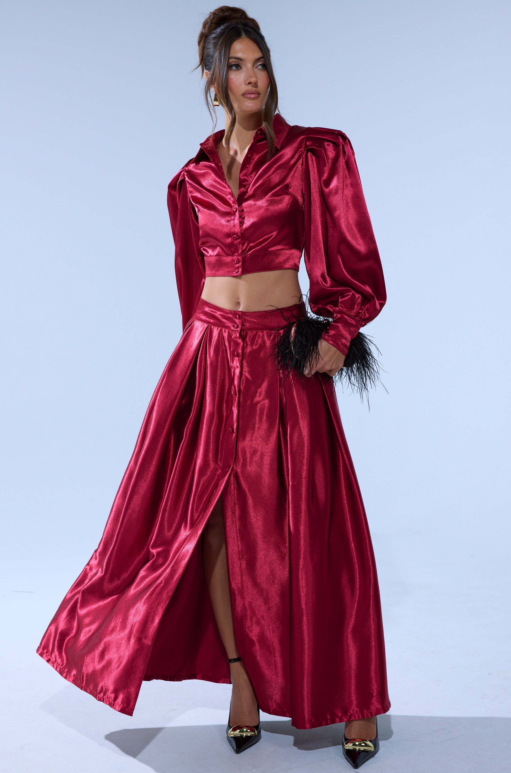 A woman poses in a shiny, deep red CANDICE OPEN BACK SATIN BUTTON DOWN SHIRT paired with a matching full-length skirt. She accessorizes with black heels and carries a small black feathered accessory.