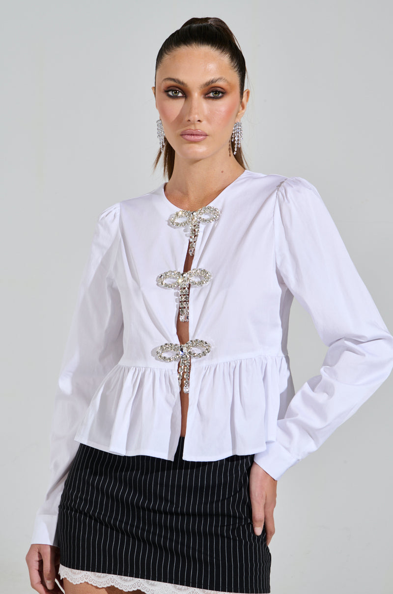 A model poses coquette-style in a DIAMOND BOW BUTTON UP, a white puff sleeve blouse with three large decorative brooches down the front, paired with a black pinstripe skirt with lace hem. Her long hair is pulled back and she's wearing sparkly earrings, standing against a plain backdrop.
