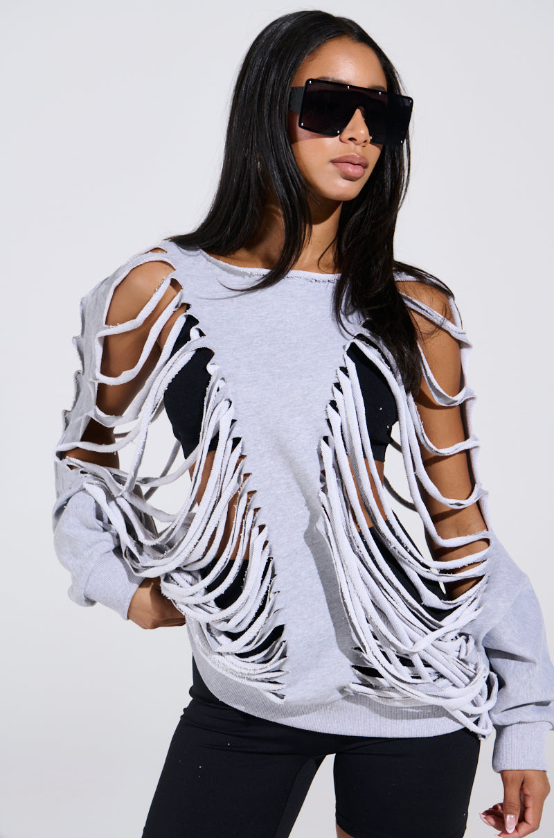 Wearing large sunglasses, a person in a CUT HIM OFF FRENCH TERRY SWEATSHIRT with distressed gray paneling reveals a black top underneath. The oversized silhouette flatters their long dark hair as they pose against a plain white background.
