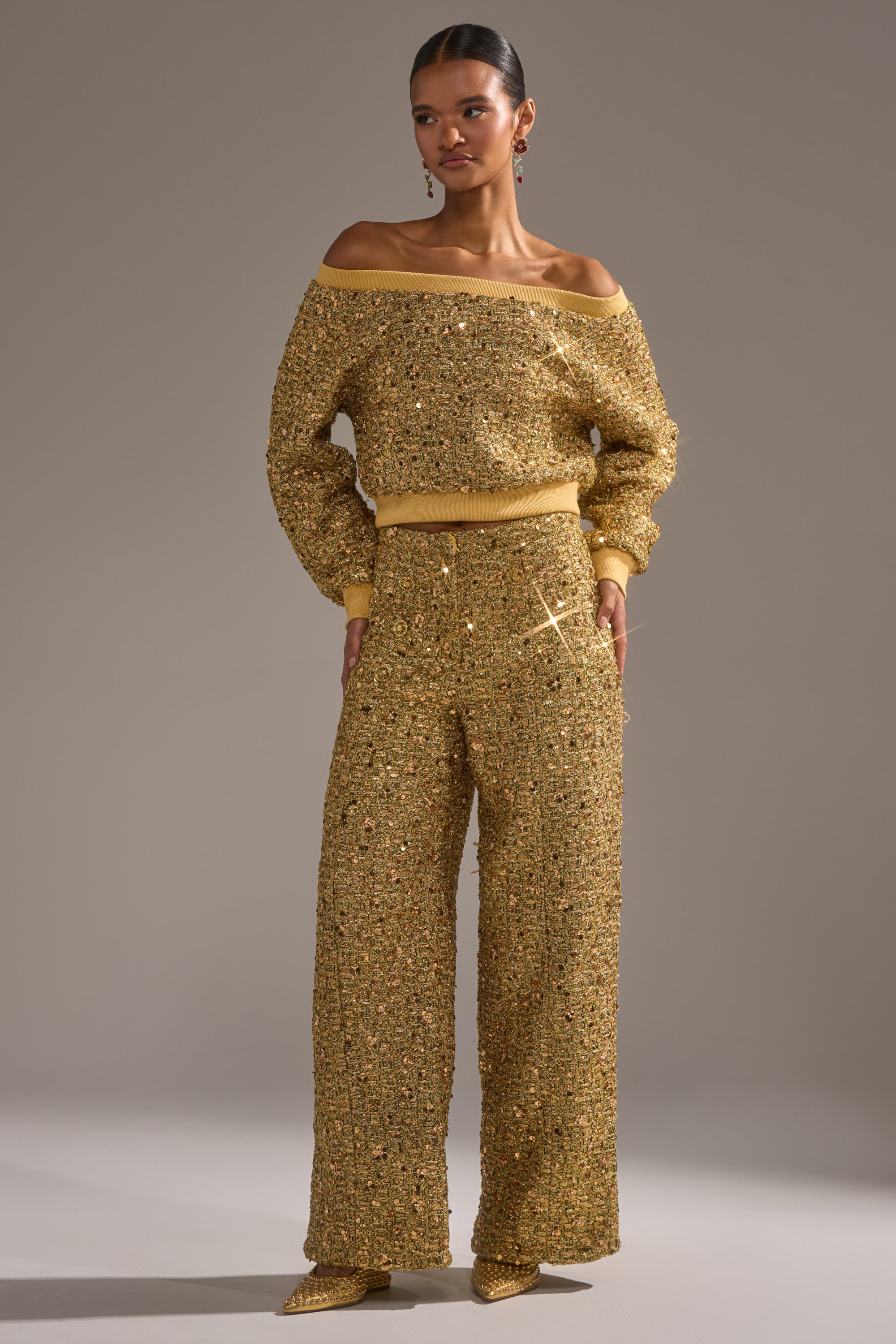 A woman stands against a plain background wearing PRETTY IN GOLD STRAIGHT LEG PANT and a matching off-the-shoulder gold sequin top, paired with pointed gold shoes.