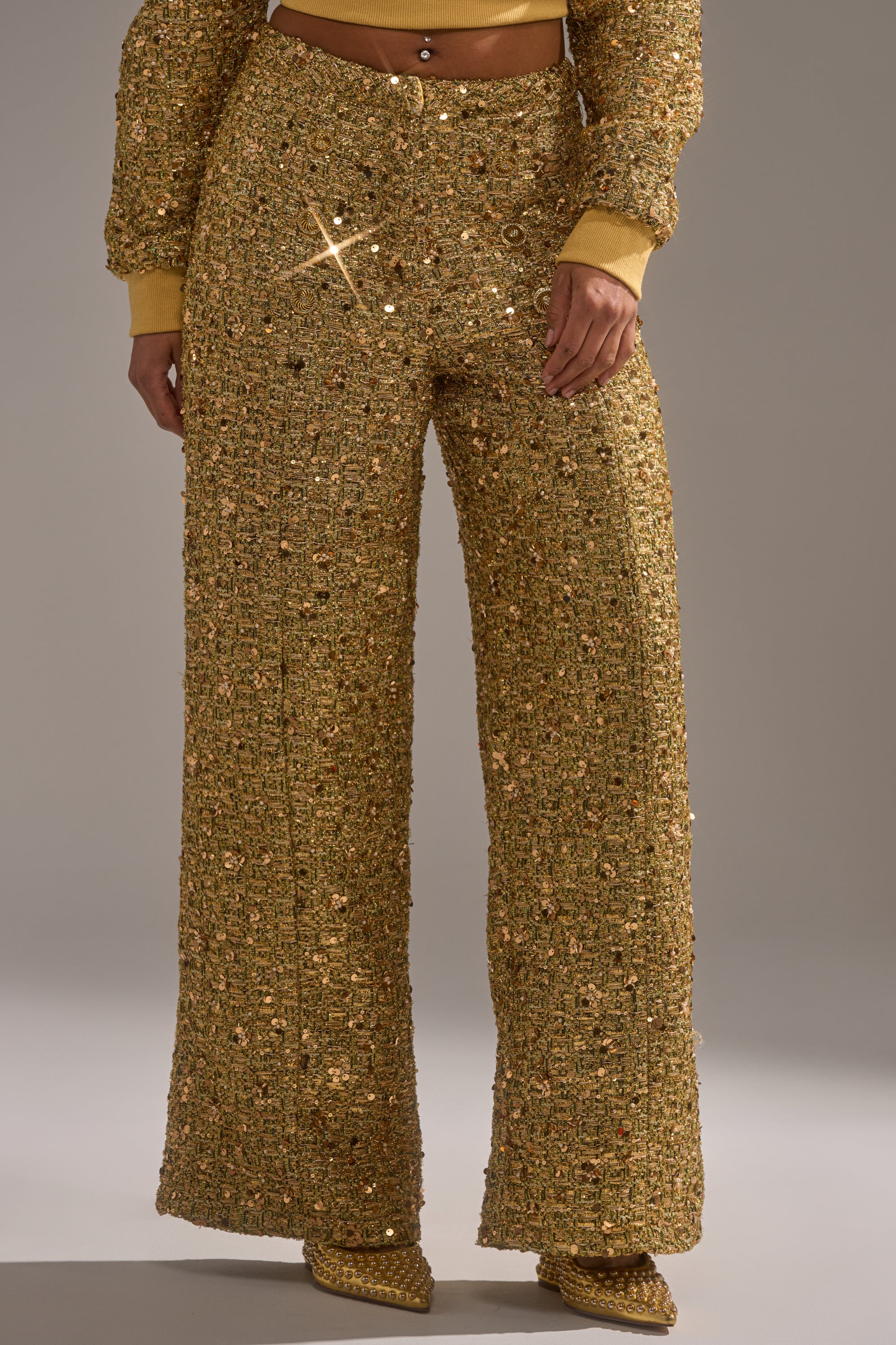 A person wearing the PRETTY IN GOLD STRAIGHT LEG PANT, featuring glittering gold sequins and a high waist, paired with pointy-toe shoes. A matching gold long-sleeve cropped top is partially visible.